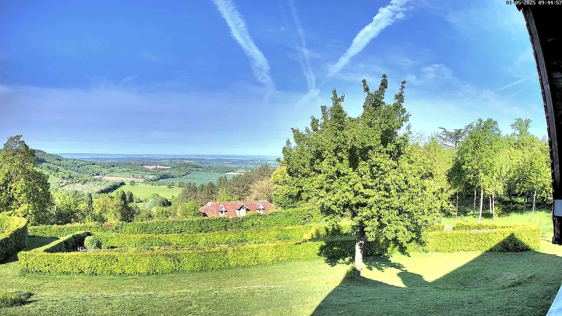 A sunny day reveals a sprawling green landscape with neatly trimmed hedges, a small house, and a distant valley under a mostly clear blue sky with contrails.