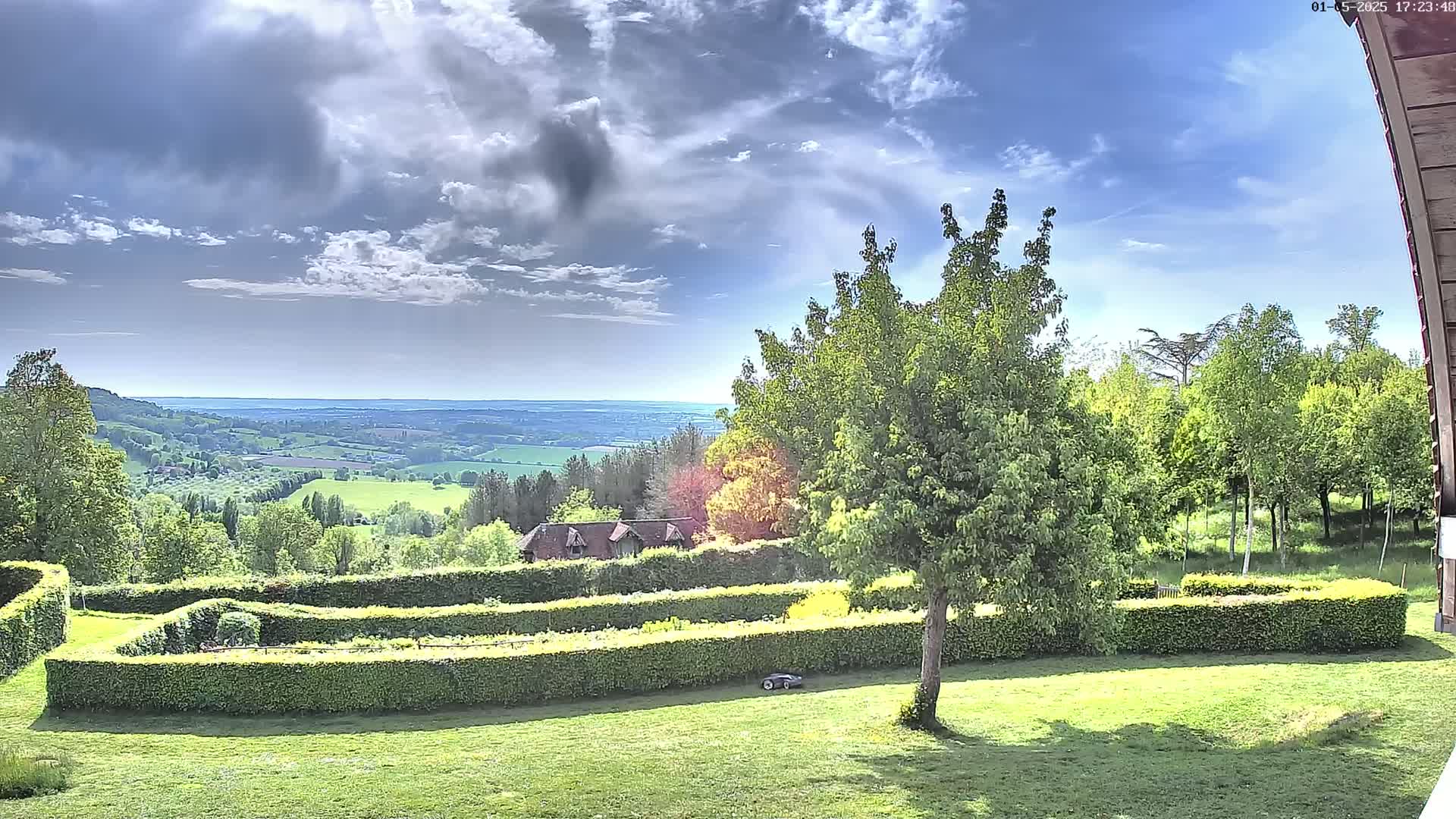 A partly cloudy sky overlooks a verdant landscape featuring a manicured lawn, hedges forming geometric patterns, a small building, and a distant rolling valley.