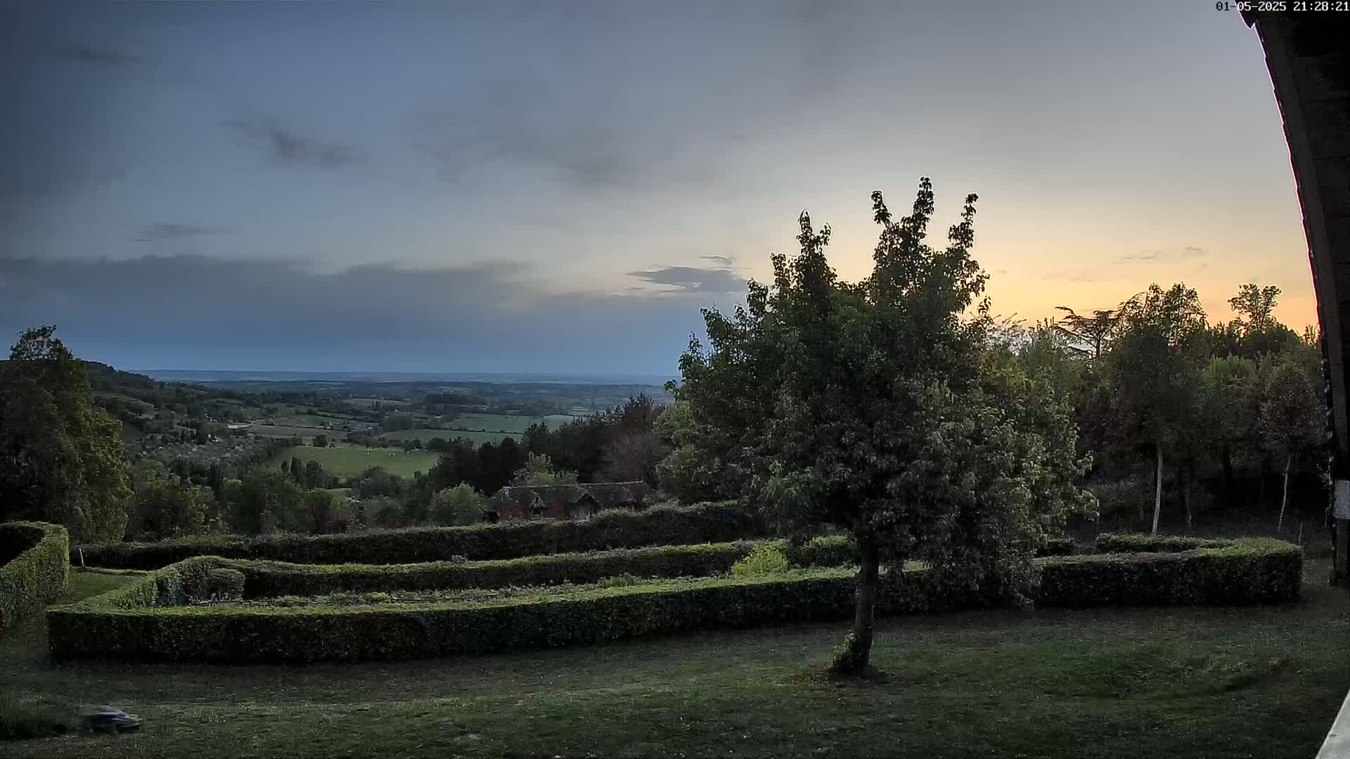 A sunset view over a rolling landscape of green fields and forests, seen from a garden with neatly trimmed hedges under a partly cloudy sky.
