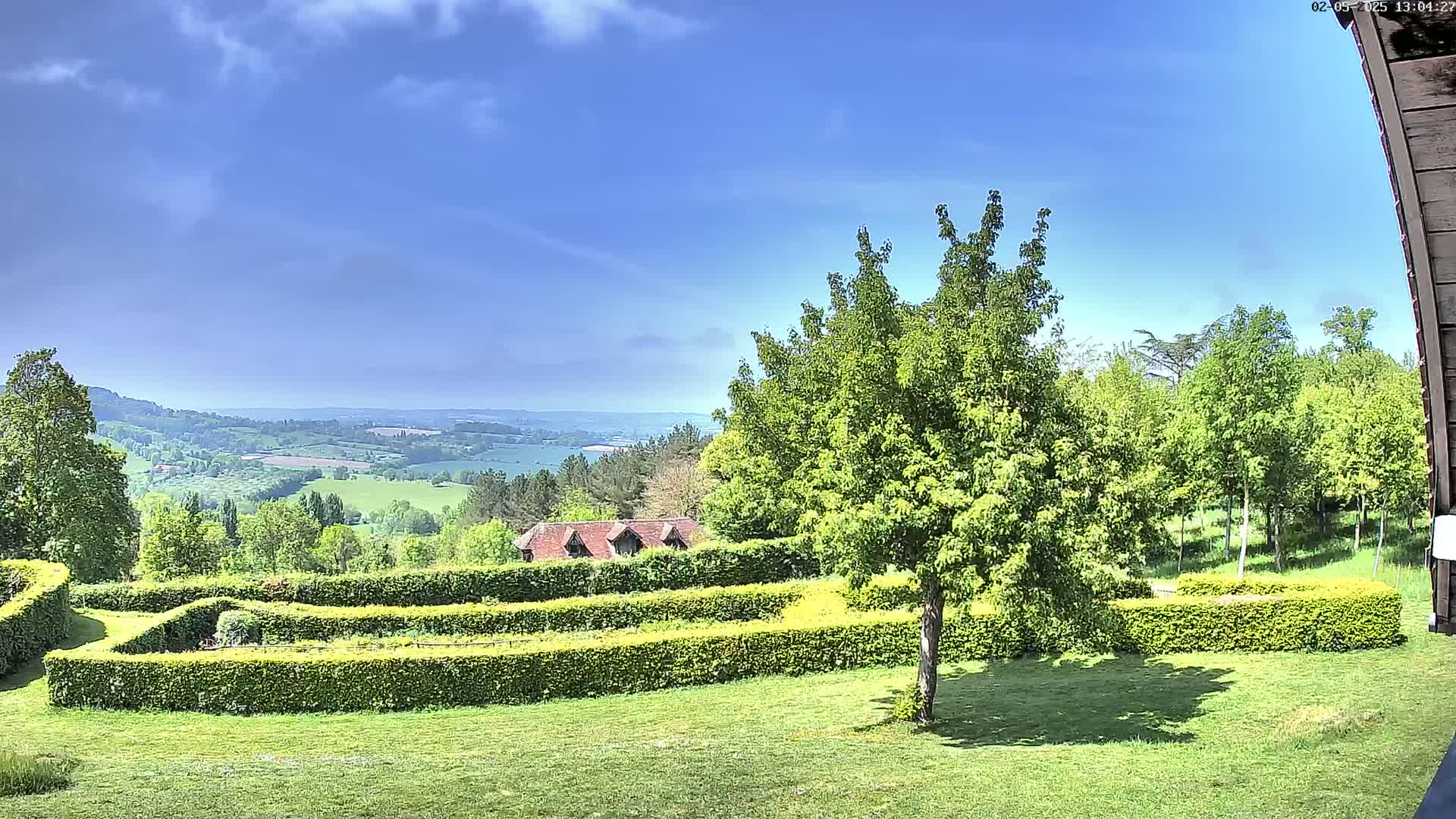 A sunny day reveals a lush green landscape with neatly trimmed hedges, a small building, and a distant view of rolling hills under a partly cloudy sky.