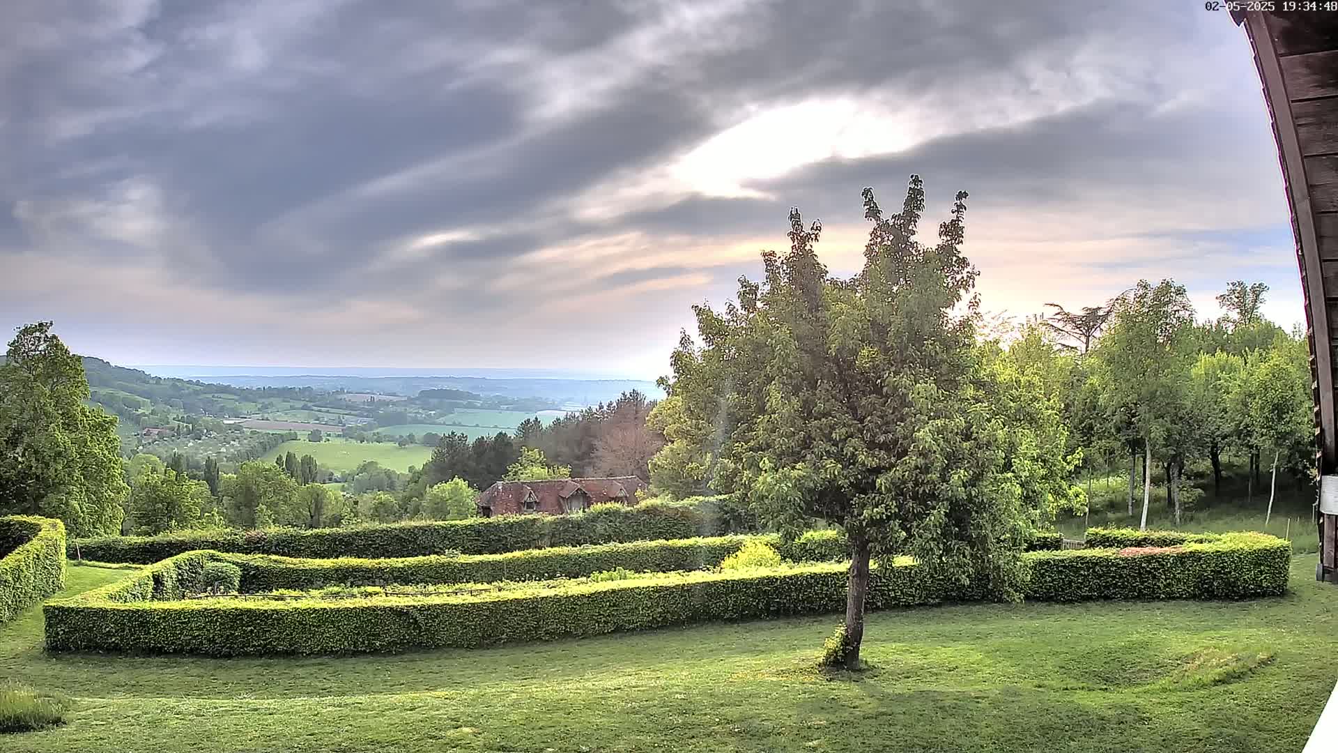 A sprawling green landscape with neatly trimmed hedges and a tree in the foreground overlooks a valley under a partly cloudy sky.