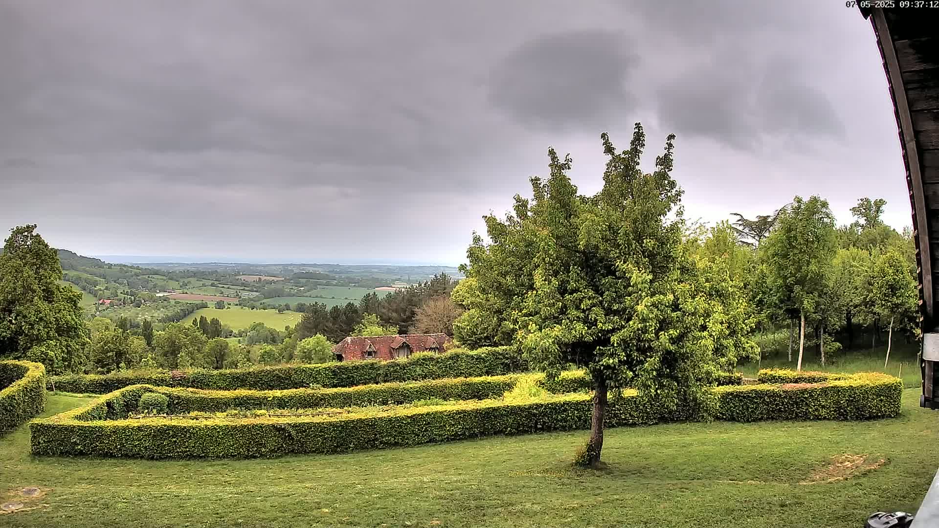 A verdant landscape featuring a manicured hedge maze overlooks a sprawling valley under a gray, overcast sky.