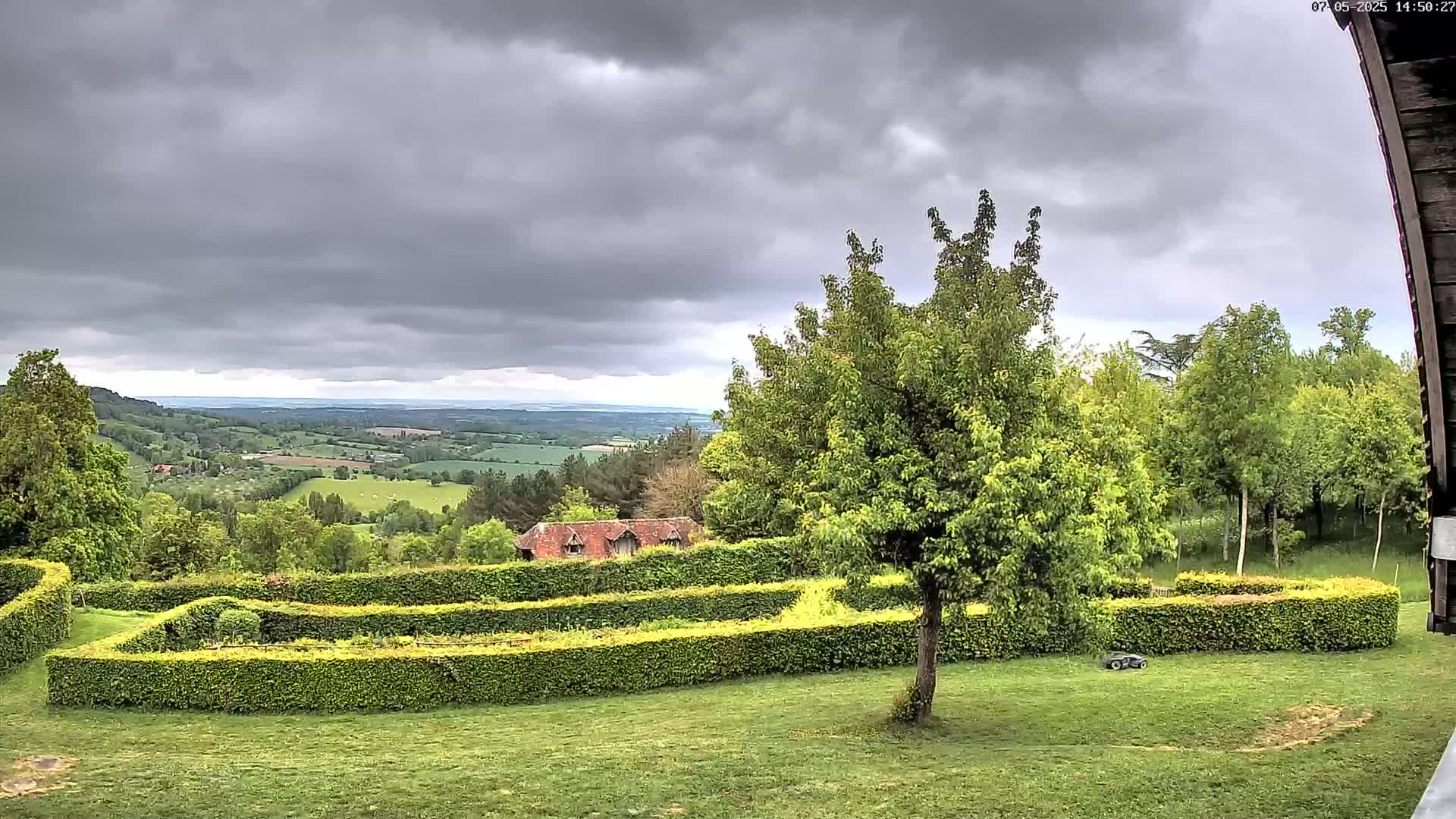 A sprawling green landscape under a cloudy sky features a geometrically-shaped hedge garden overlooking a distant valley.