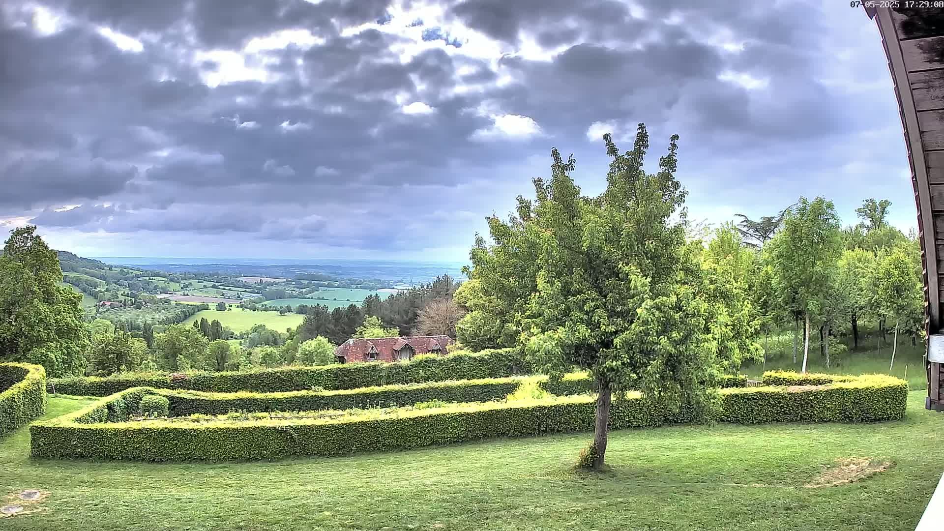 A meticulously manicured garden with boxwood hedges overlooks a rolling green valley under a cloudy sky.