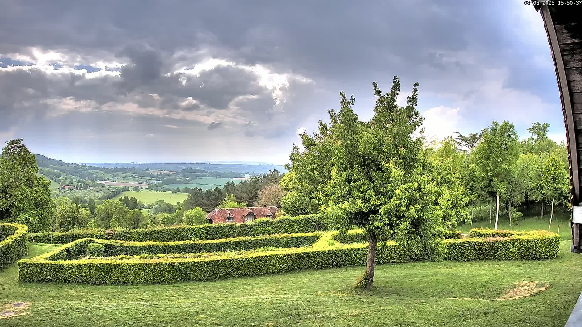 A verdant landscape, featuring a meticulously manicured hedge maze, overlooks a rolling countryside under a partly cloudy sky.