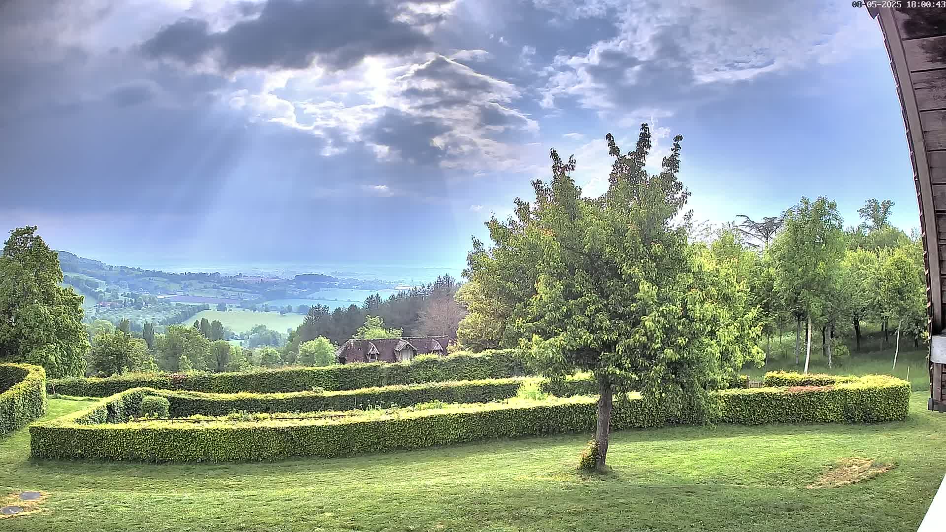 A meticulously manicured garden with hedges, overlooking a sprawling countryside under a partly cloudy sky.