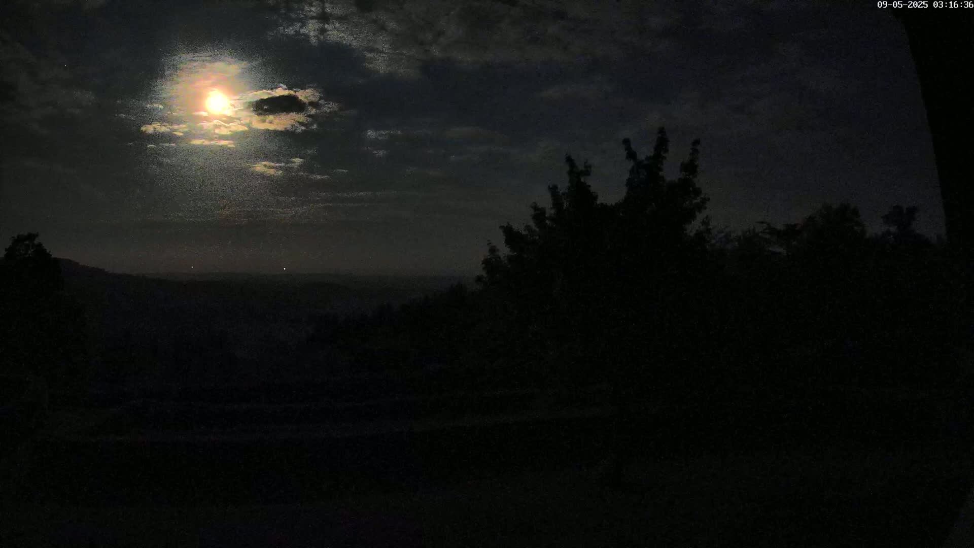 A bright moon shines through partly cloudy night skies over a dark, silhouetted landscape of trees and hills.