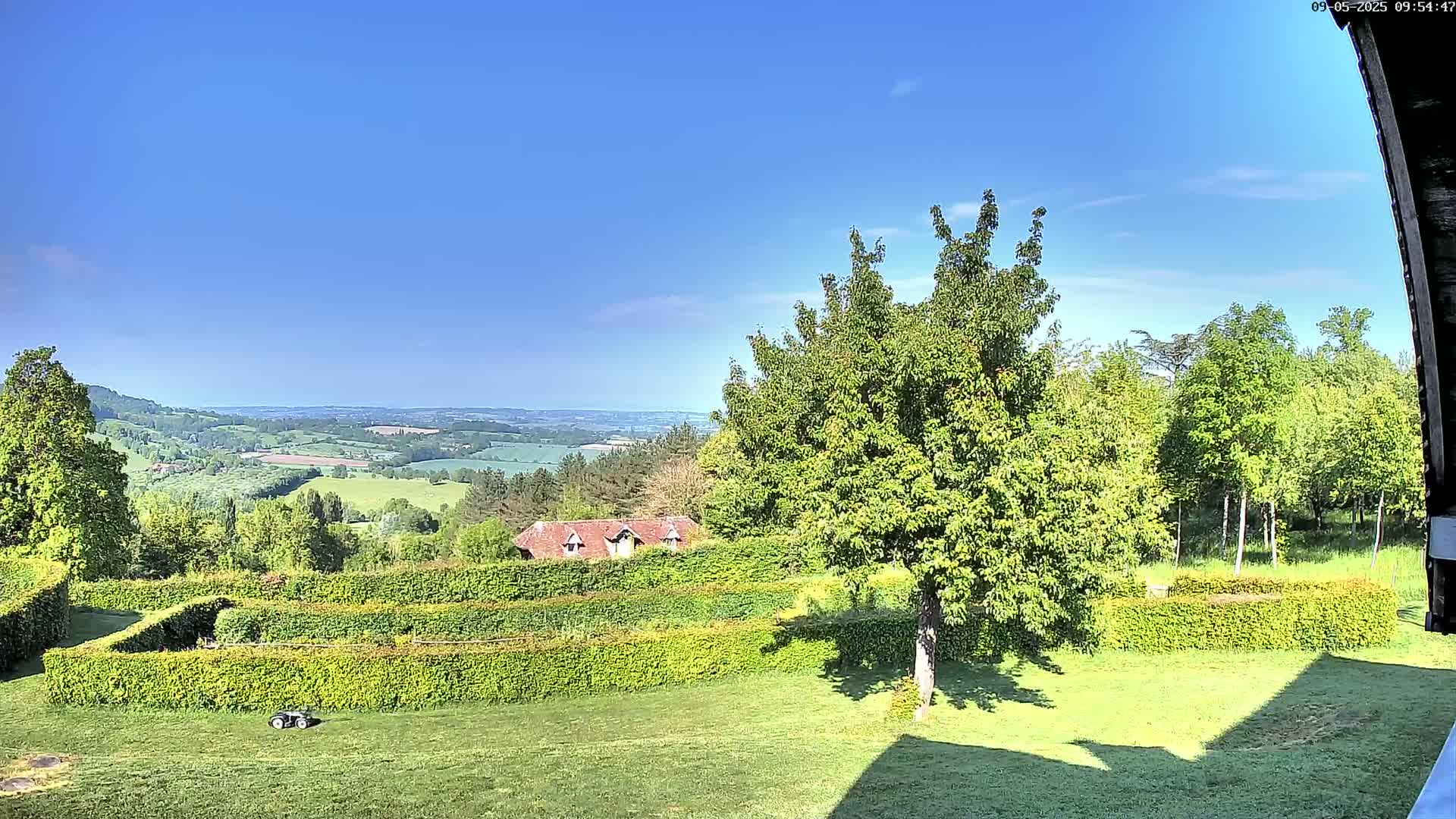 A sunny day reveals a verdant landscape featuring a manicured lawn, neatly trimmed hedges forming geometric patterns, a small house nestled amongst trees, and a distant view of rolling hills under a clear blue sky.