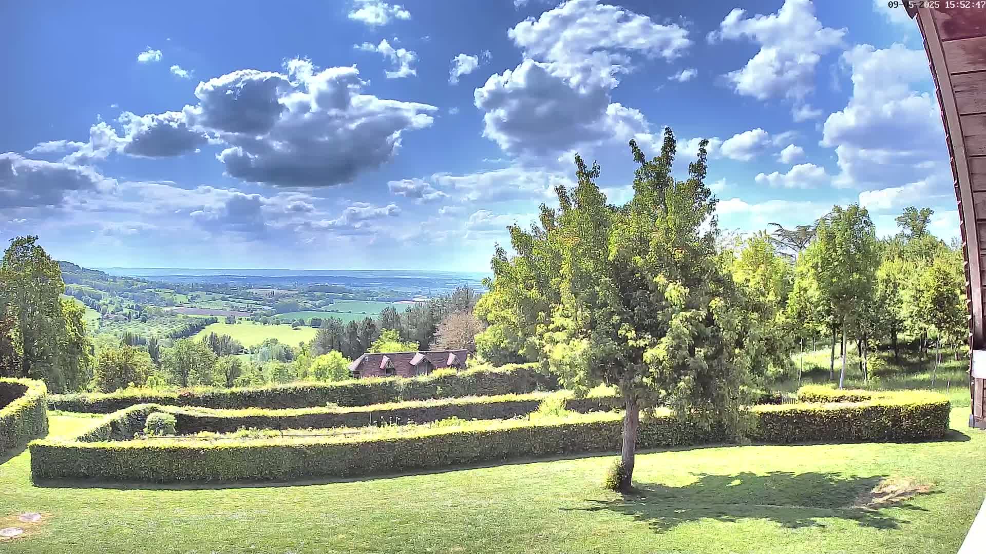 A partly sunny day reveals a meticulously landscaped garden with a neatly trimmed hedge maze, overlooking a sprawling green valley under a partly cloudy sky.