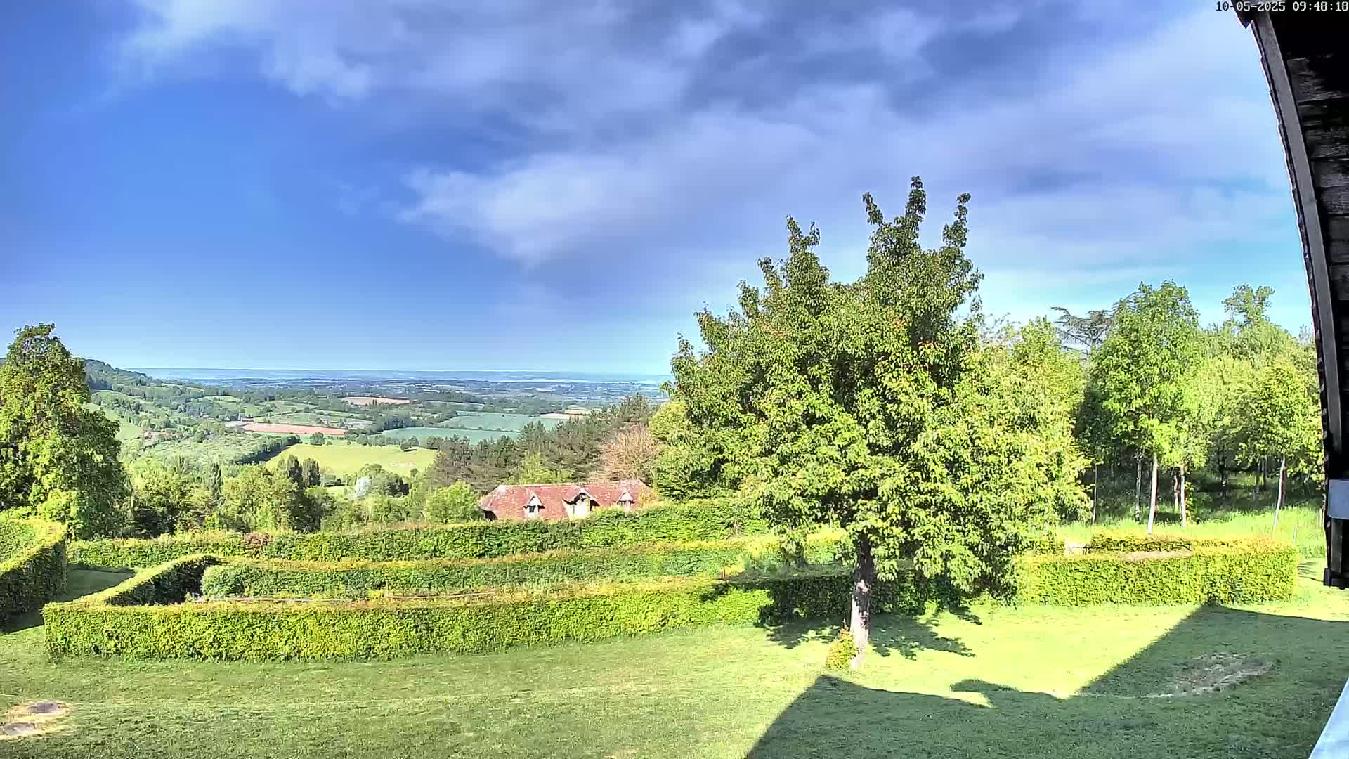 A partly cloudy sky overlooks a sprawling green landscape with neatly trimmed hedges, a small building, and distant hills, culminating in a hazy horizon.