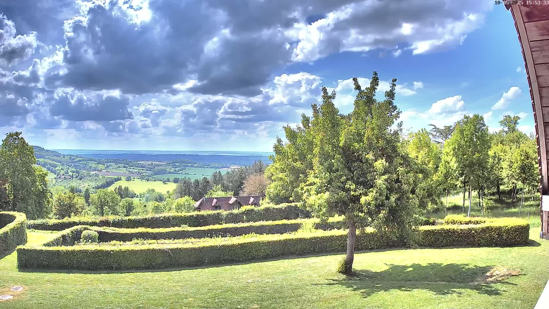 A partly cloudy sky overlooks a verdant landscape featuring a manicured garden, a small building, and a rolling expanse of farmland in the distance.