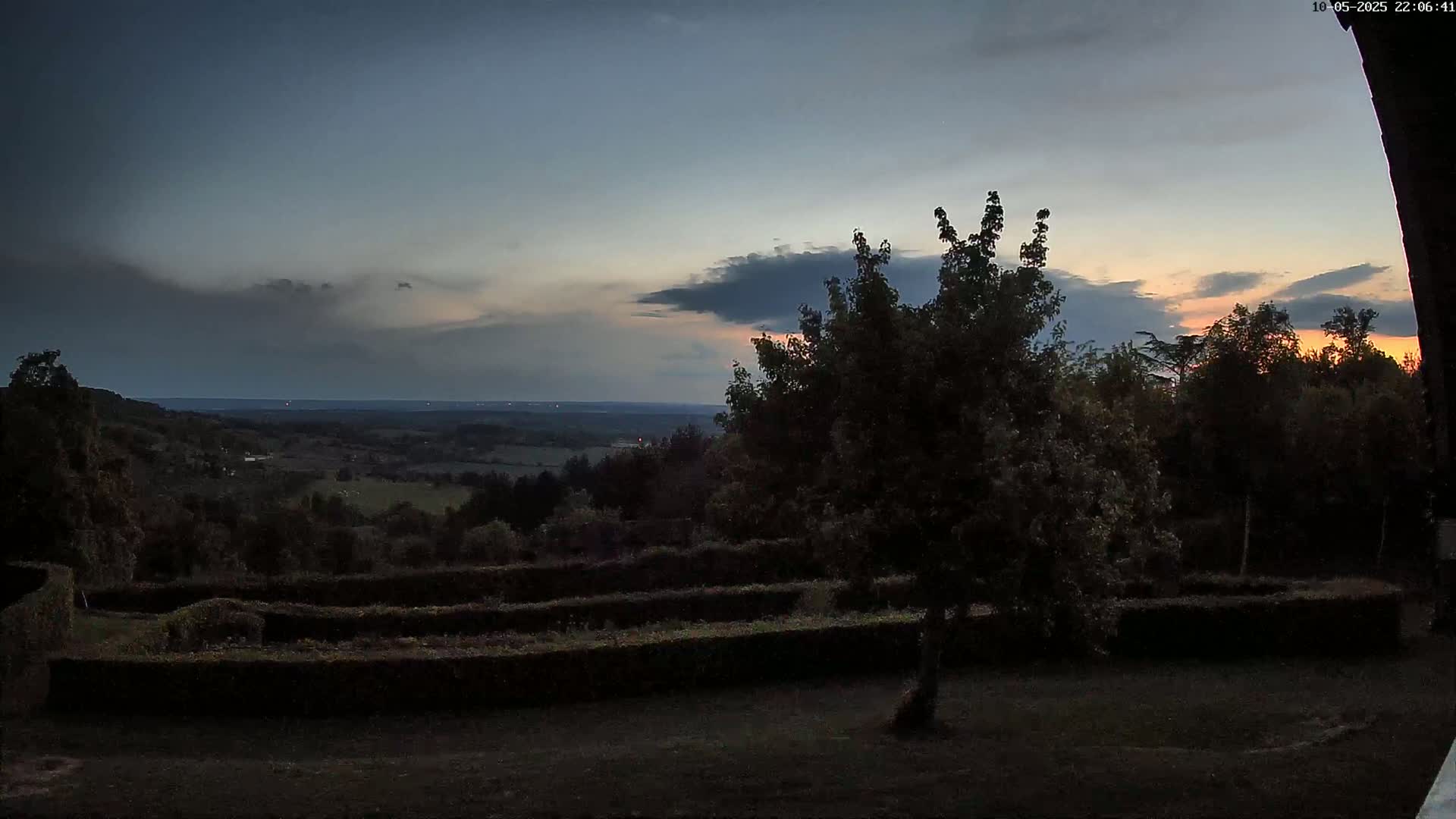 A twilight view of a hilly landscape, showing a distant horizon under a mostly cloudy sky with some hints of orange at sunset, overlooked by a garden with neatly trimmed hedges.