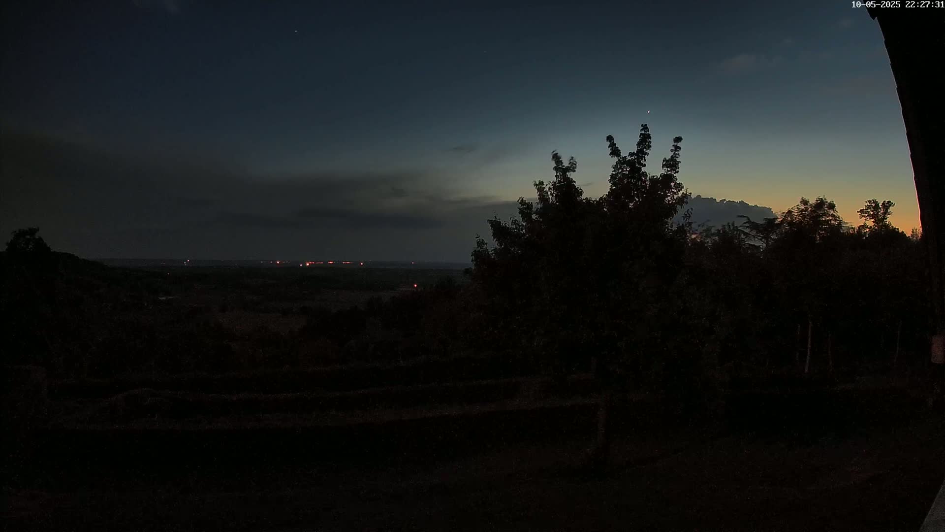 A nighttime view of a dark landscape with a few distant lights, silhouetted trees in the foreground, and a partly cloudy sky showing hints of twilight.