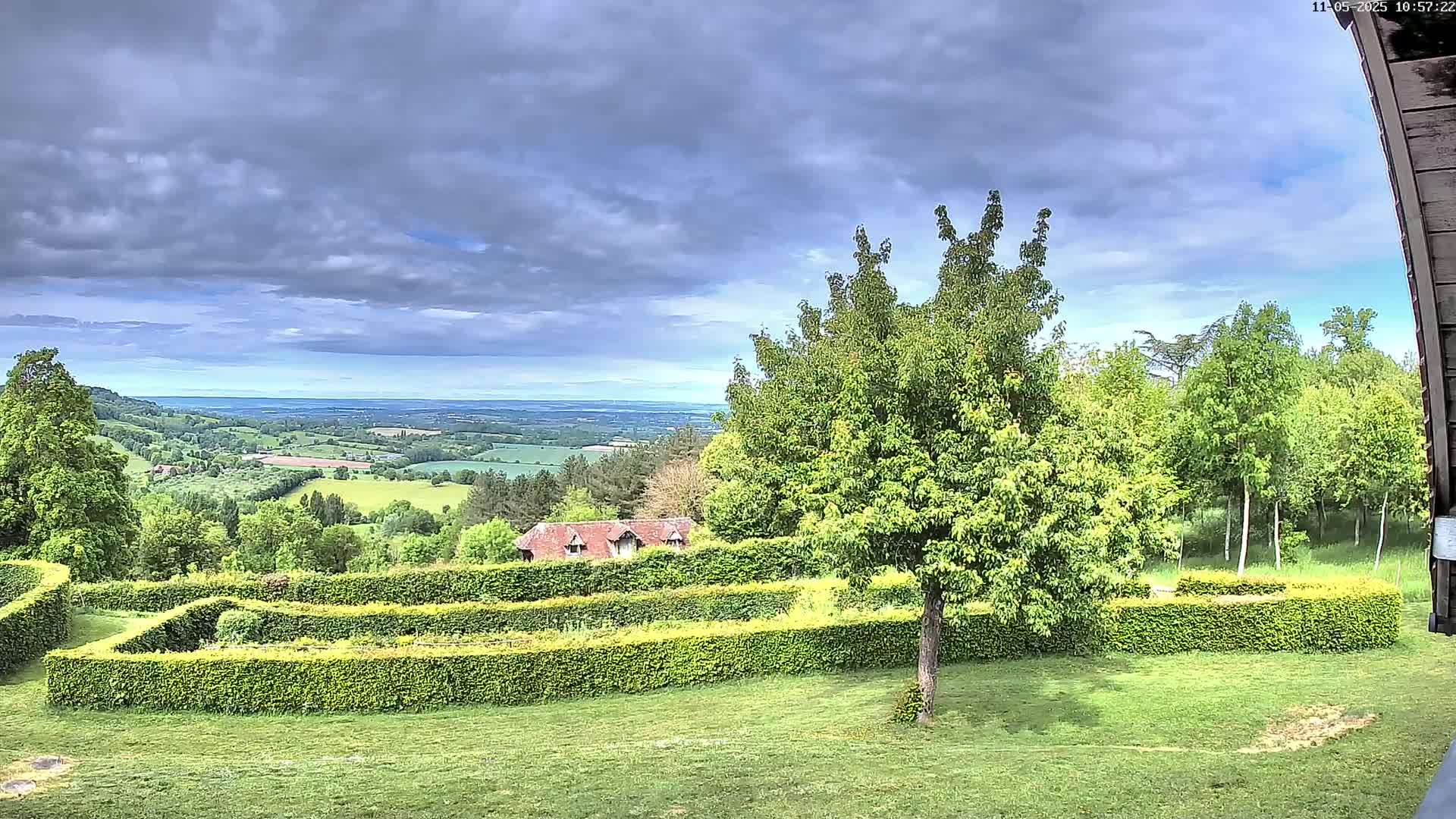 A lush green garden with neatly trimmed hedges overlooks a rolling landscape under a partly cloudy sky.