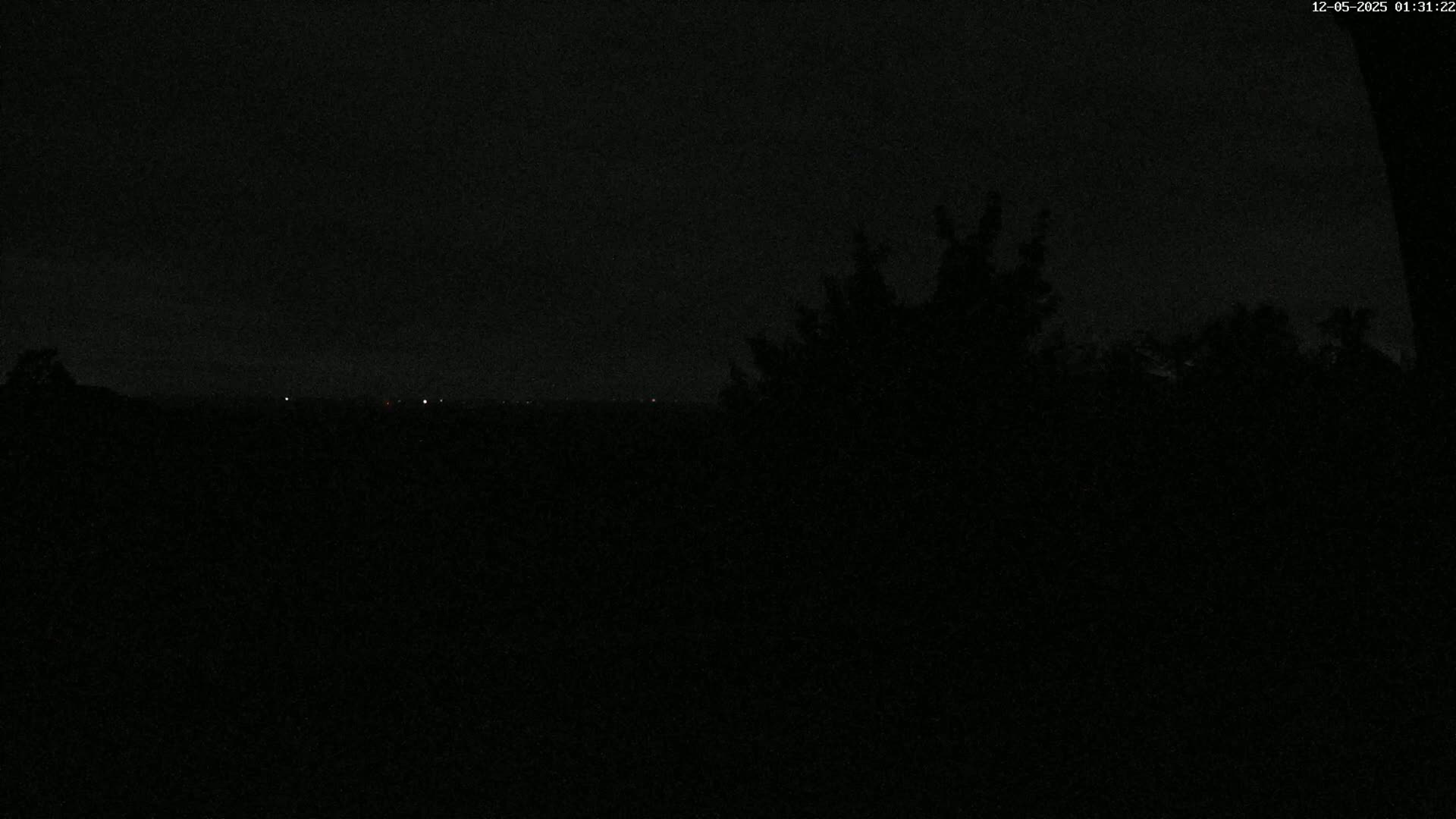 A nighttime view shows a dark landscape with a few distant lights and silhouetted trees in the foreground under a dark sky.