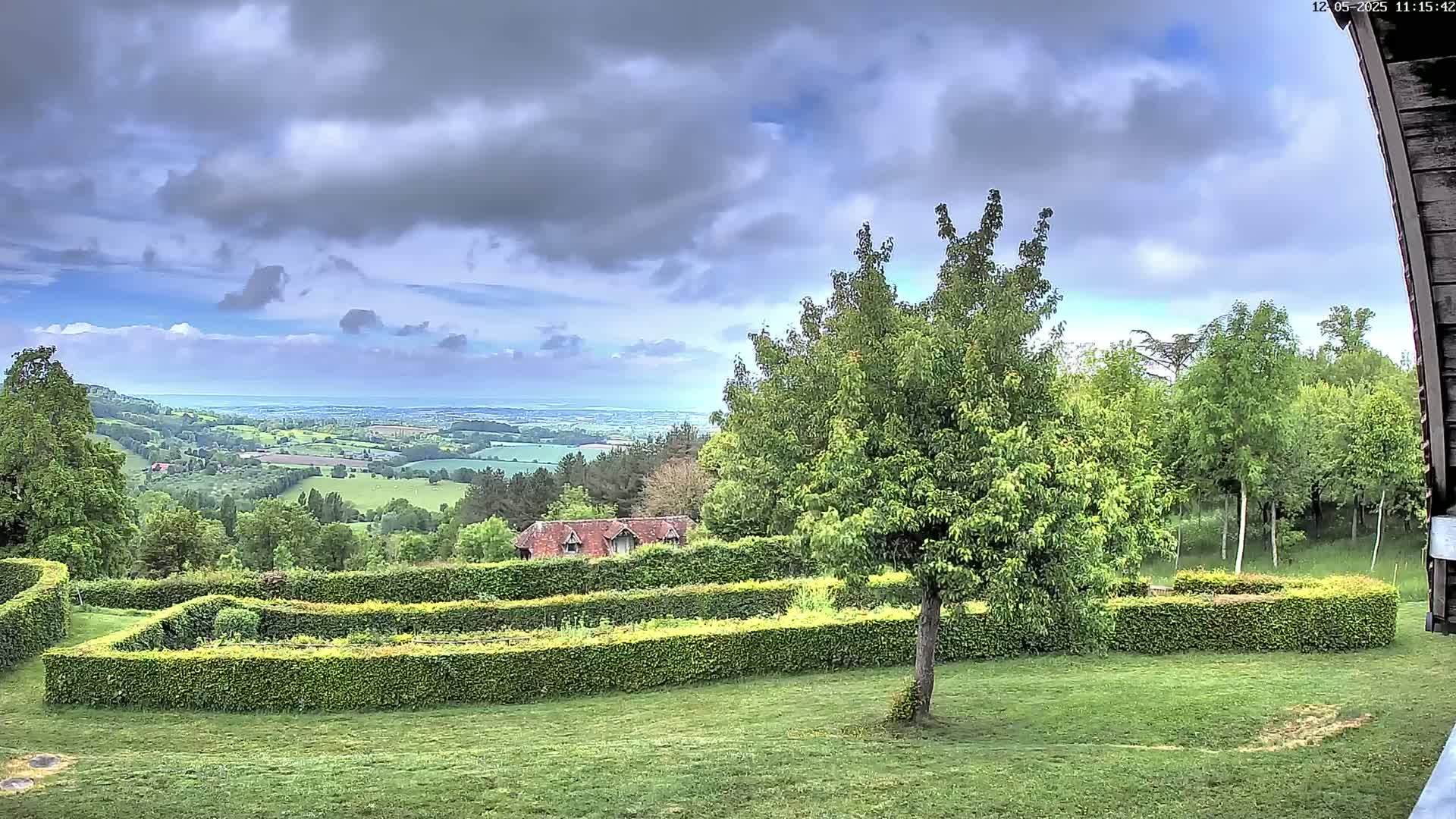 A verdant landscape featuring a meticulously manicured hedge garden, a small building, and a sprawling view of a valley under a partly cloudy sky.
