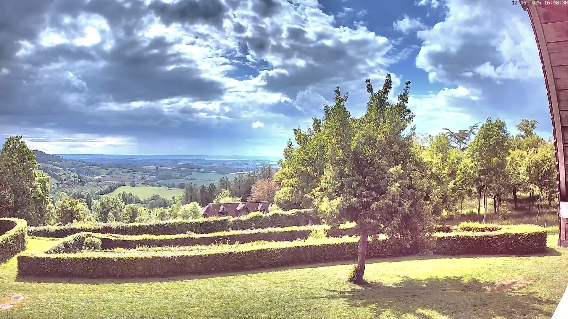 A partly cloudy sky overlooks a verdant landscape featuring a manicured garden with hedges, trees, and a distant view of rolling hills.