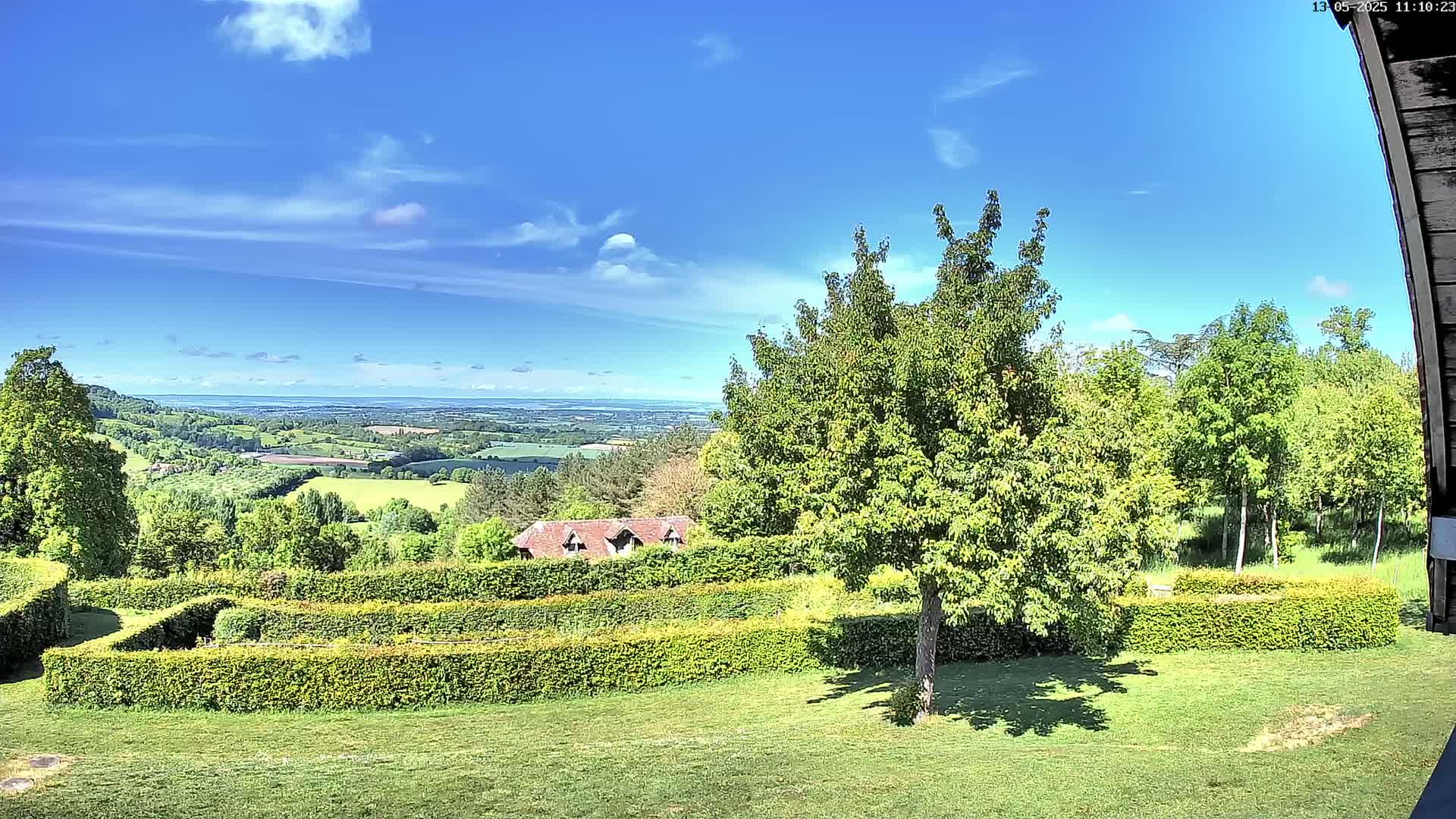 A sunny day reveals a sprawling landscape of green fields and trees, a distant body of water, and a neatly manicured garden with a small building nestled among the foliage.