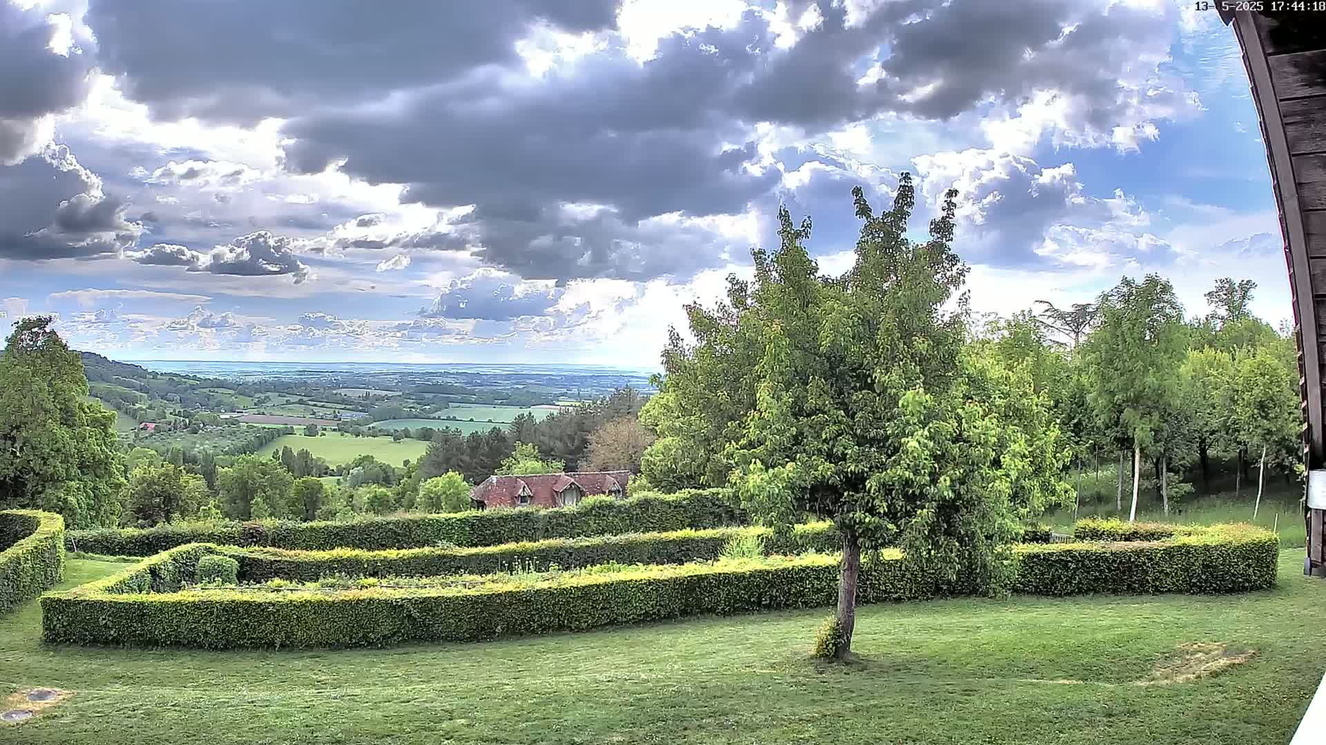 A partly cloudy sky overlooks a verdant landscape featuring a meticulously manicured hedge garden, several trees, and a distant view of rolling hills and a body of water.