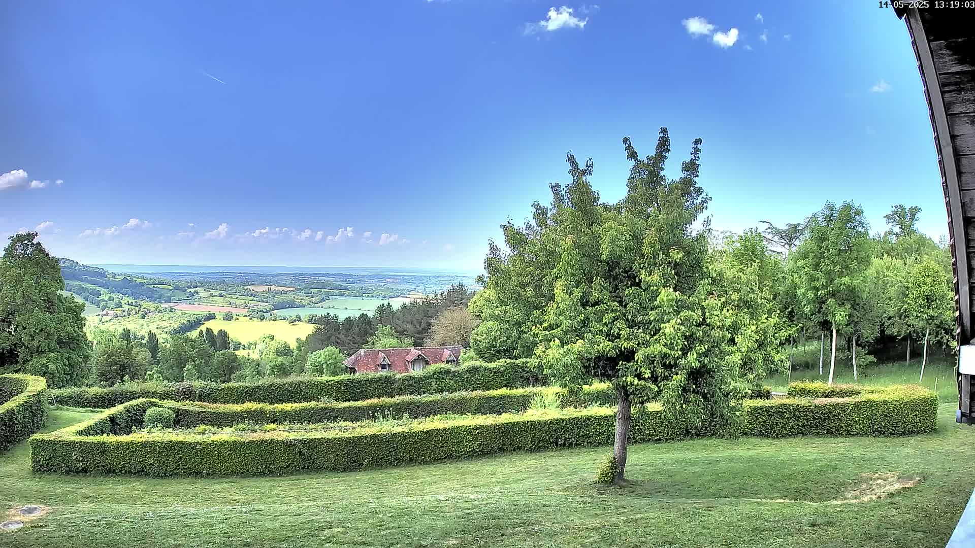 A verdant landscape with neatly trimmed hedges, a small building, and various trees stretches toward a distant view of rolling hills and a clear blue sky with a few clouds.