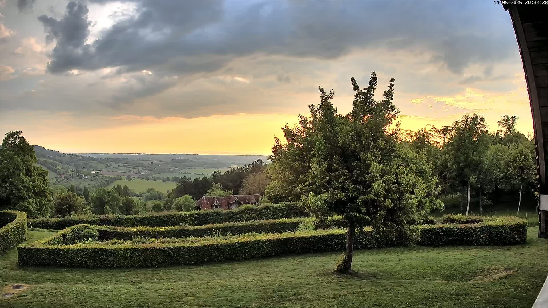 A sunset over a sprawling, verdant valley is viewed from a grassy area with a neatly trimmed hedge and a lone tree in the foreground, under a partly cloudy sky.