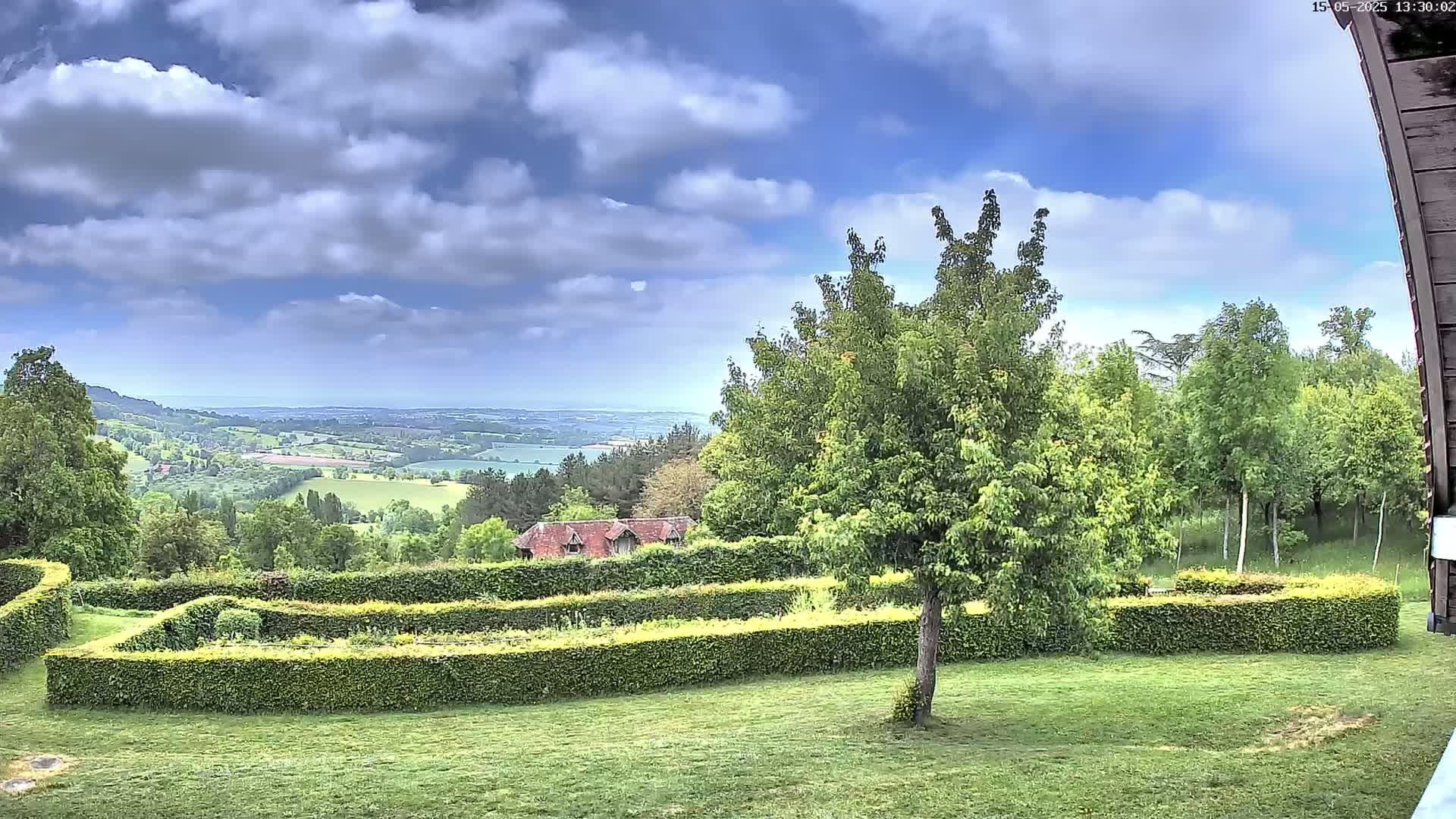 A partly cloudy sky overlooks a lush green landscape with neatly trimmed hedges, a small building, and a distant view of rolling hills.