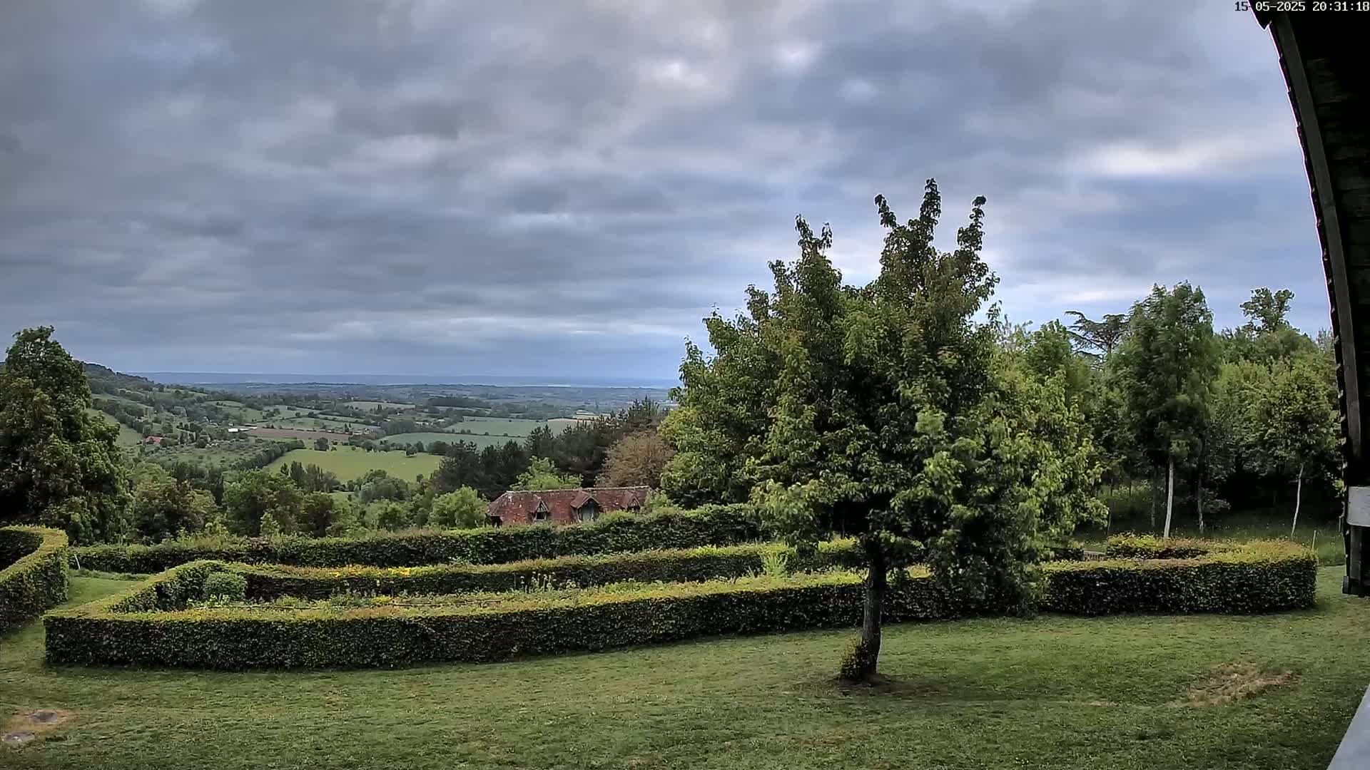 A verdant landscape unfolds under a cloudy sky, featuring a manicured garden with hedges and a distant view of rolling hills and a flat expanse.