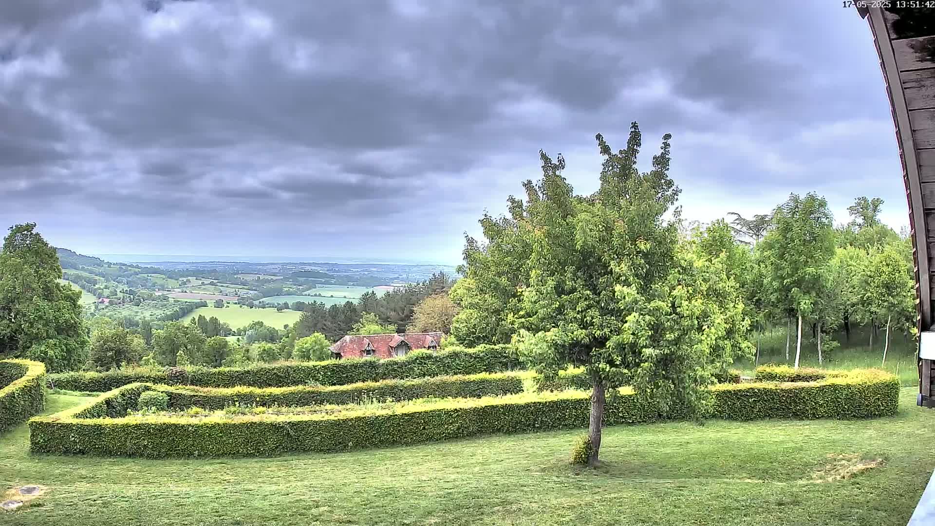 A verdant landscape with a meticulously manicured hedge maze, several trees, a small building, and a distant view of rolling hills under a cloudy sky.