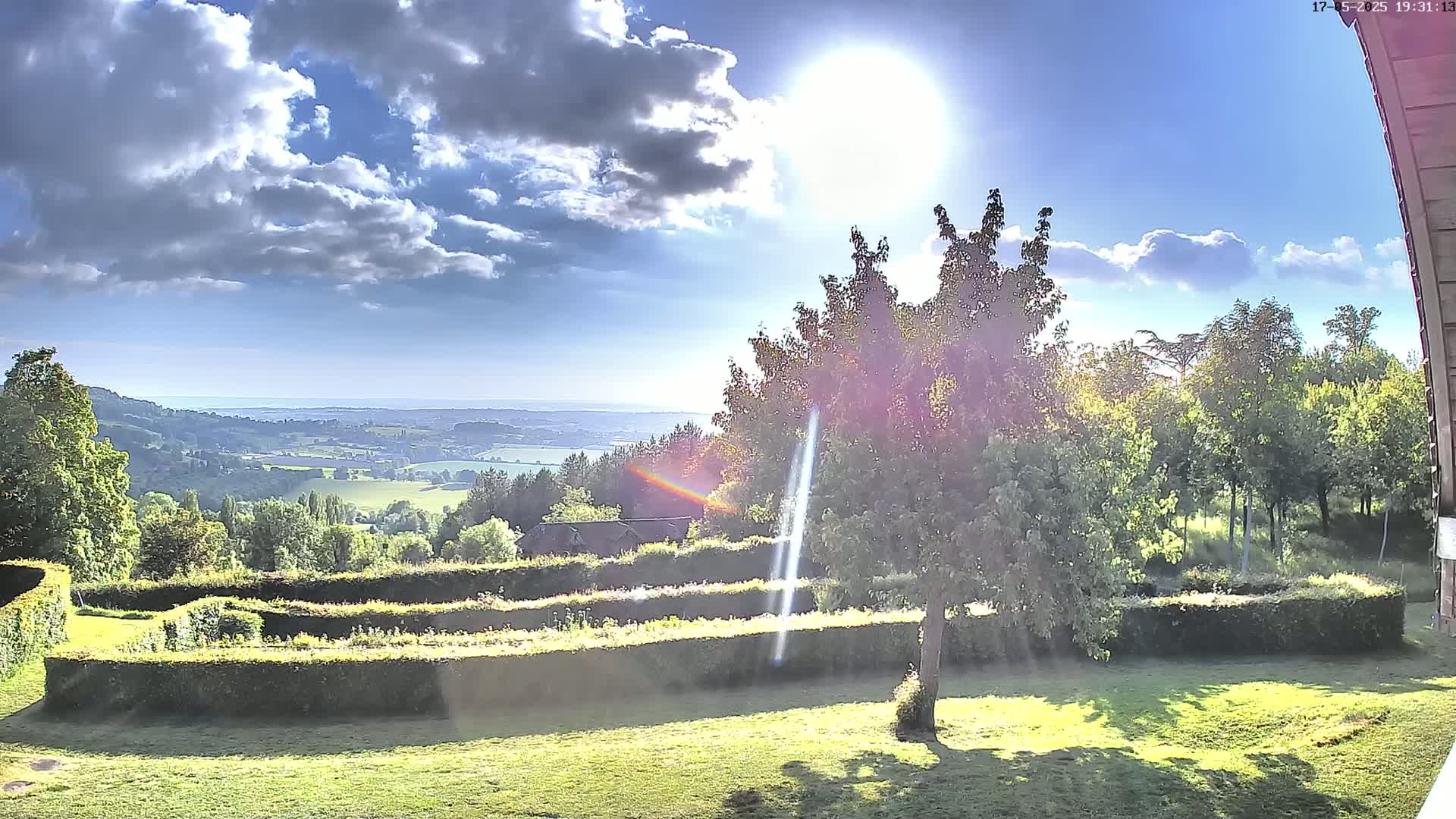 A sunny day with partly cloudy skies overlooks a verdant valley and distant sea, viewed from a garden with neatly trimmed hedges and a tree in the foreground.