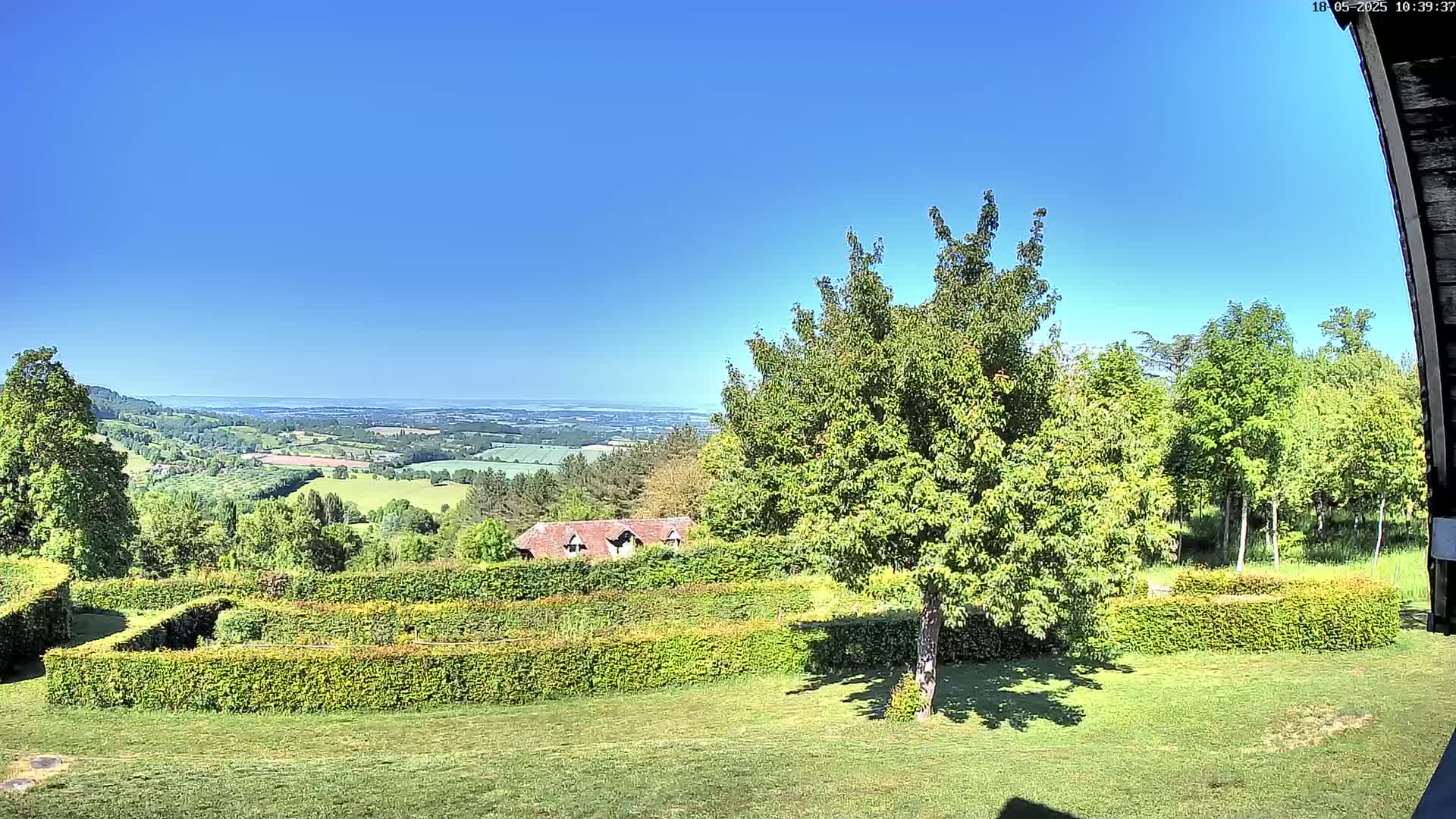 A sprawling green landscape with neatly trimmed hedges, a small building, and a distant view of rolling hills under a clear blue sky.