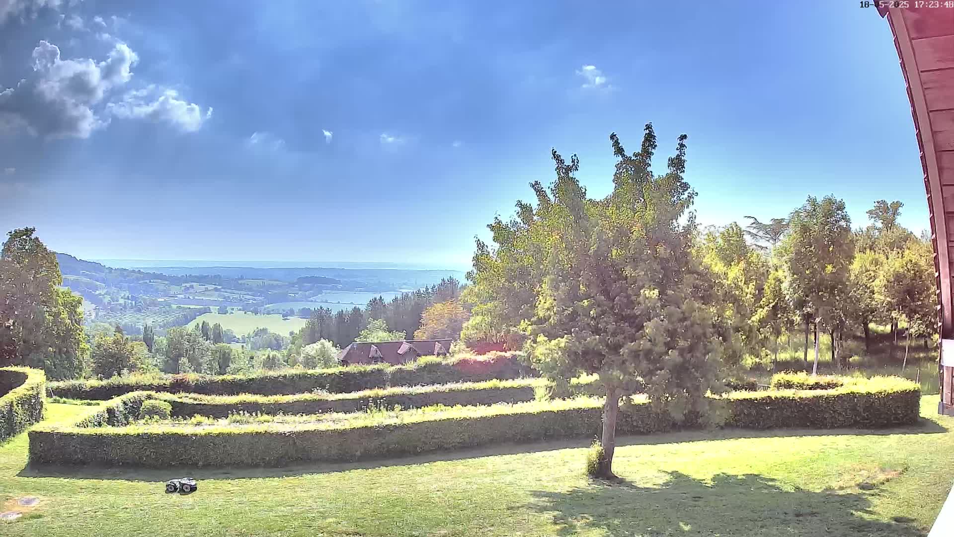 A sunny day reveals a vast, rolling landscape with a distant body of water, viewed from a manicured lawn featuring a geometric hedge design and a robotic lawnmower.