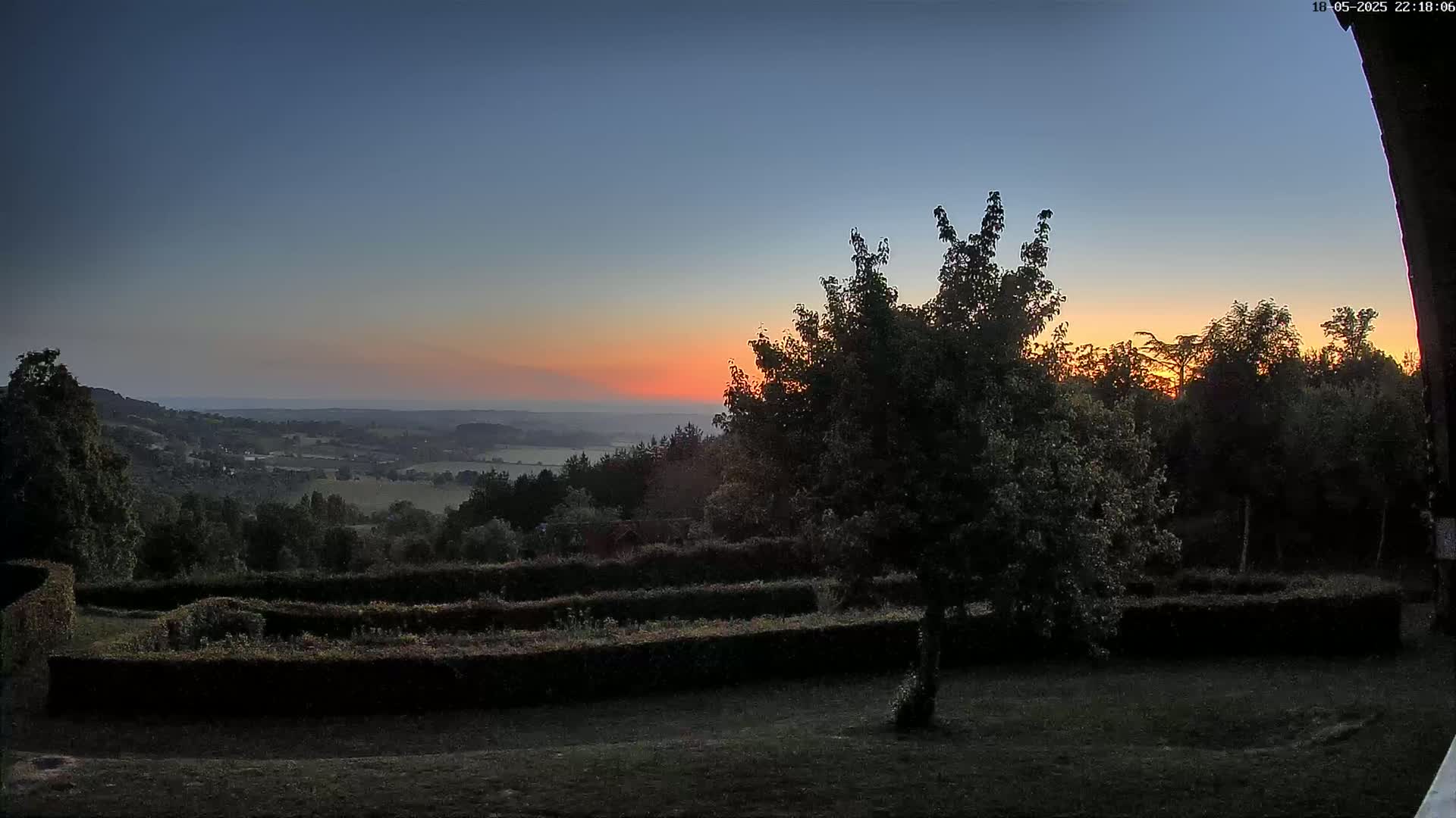 A sunset over a hilly landscape viewed from a garden with neatly trimmed hedges, under a clear sky.