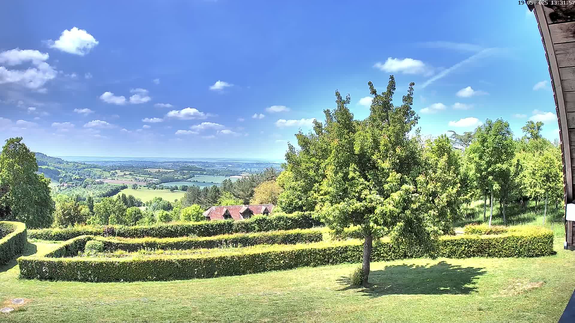 A sunny day reveals a sprawling green landscape with a neatly manicured garden featuring hedges, a small building, and a distant view of rolling hills under a partly cloudy sky.