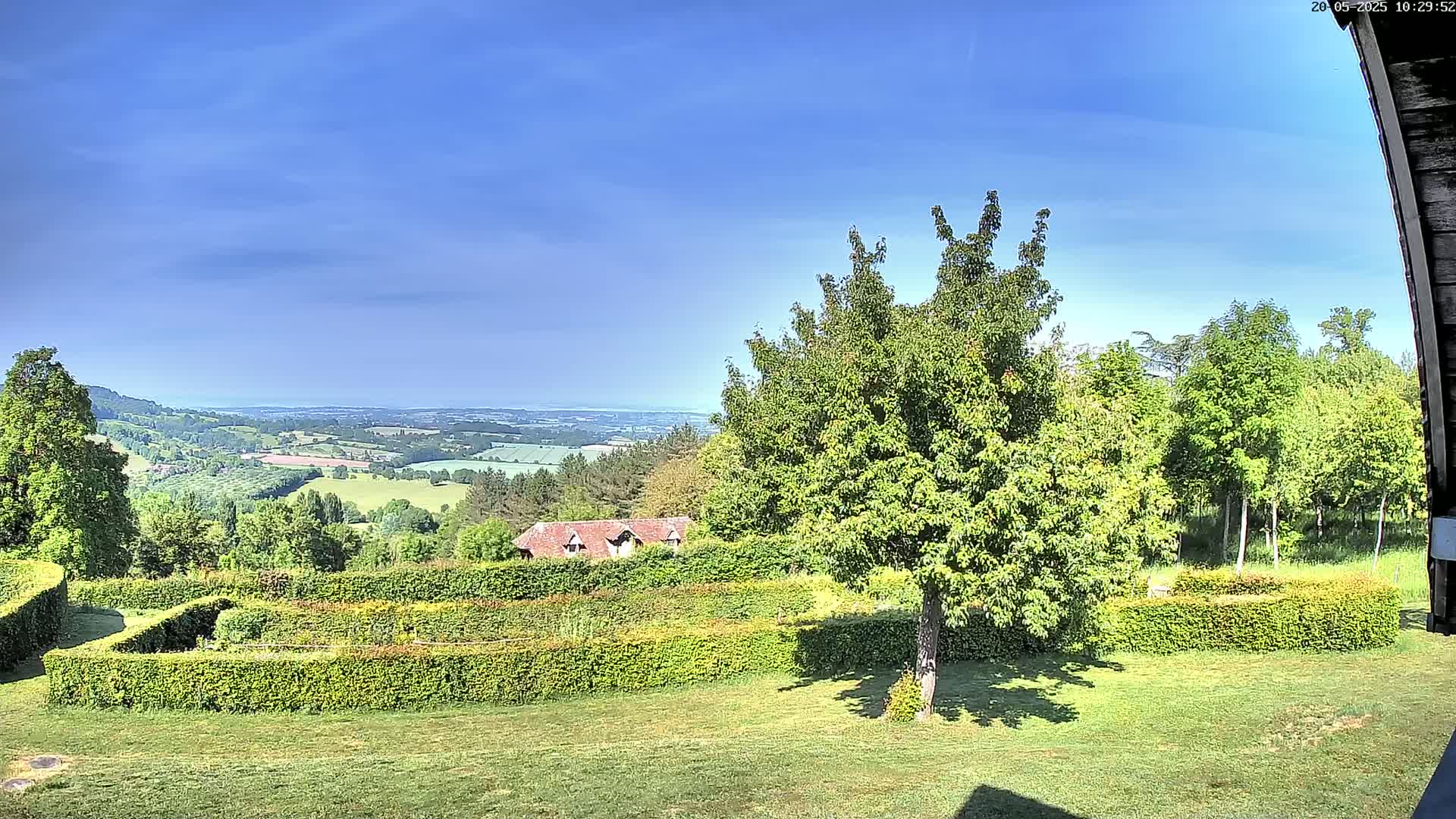 A sprawling green landscape with a neatly manicured hedge maze, a small building, and numerous trees under a clear blue sky.