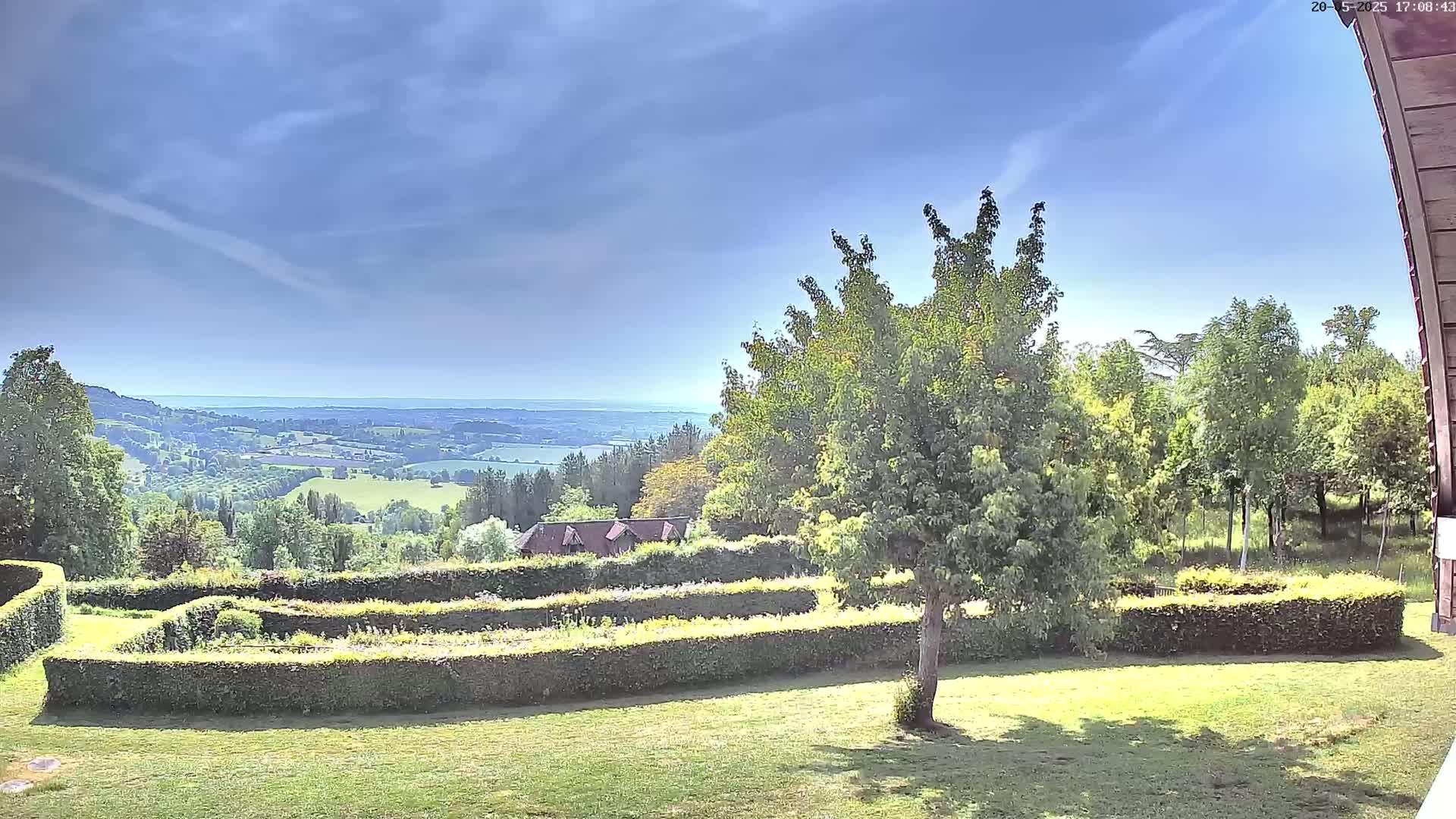 A verdant hillside garden with neatly trimmed hedges overlooks a sprawling valley and distant body of water under a mostly sunny sky.