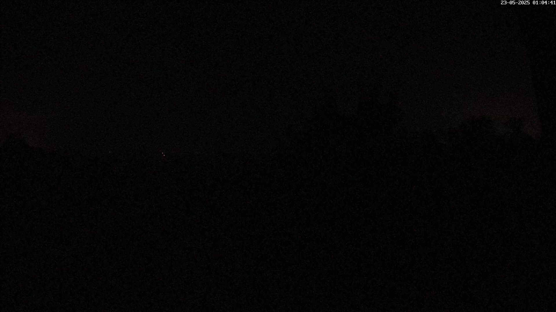 A very dark nighttime scene shows a barely visible treeline in the distance under a mostly clear, dark sky.