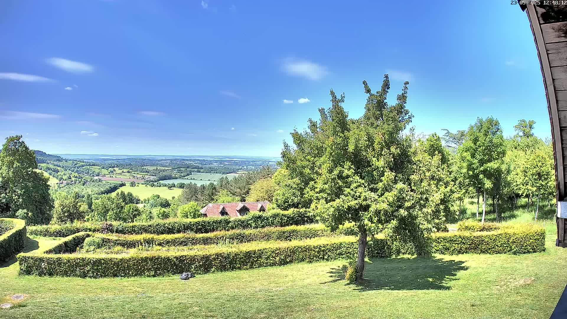 A sprawling green landscape under a bright blue sky with a few clouds, featuring a meticulously manicured garden with hedges, a small building, and a distant view of rolling hills.