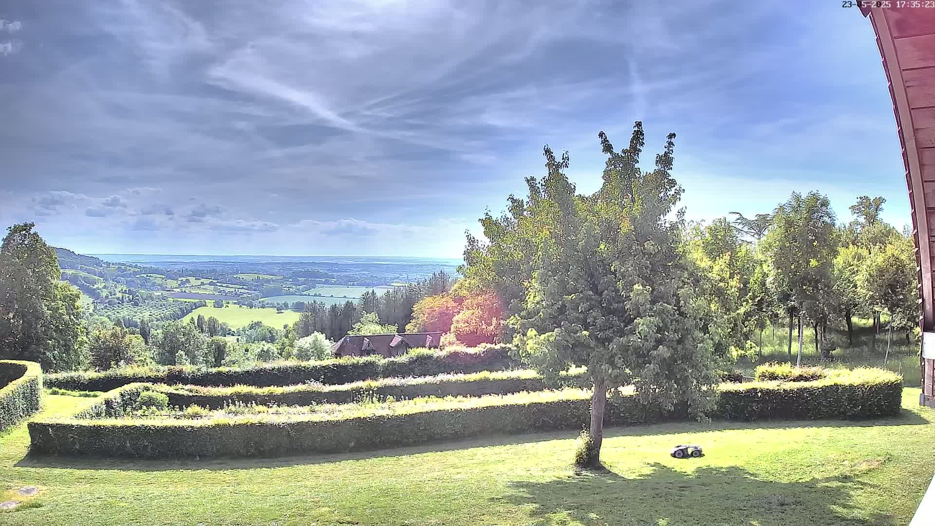 A partly sunny day reveals a sprawling green landscape with a manicured lawn, hedges forming geometric patterns, and a distant view of rolling hills and a body of water under a mostly clear sky.