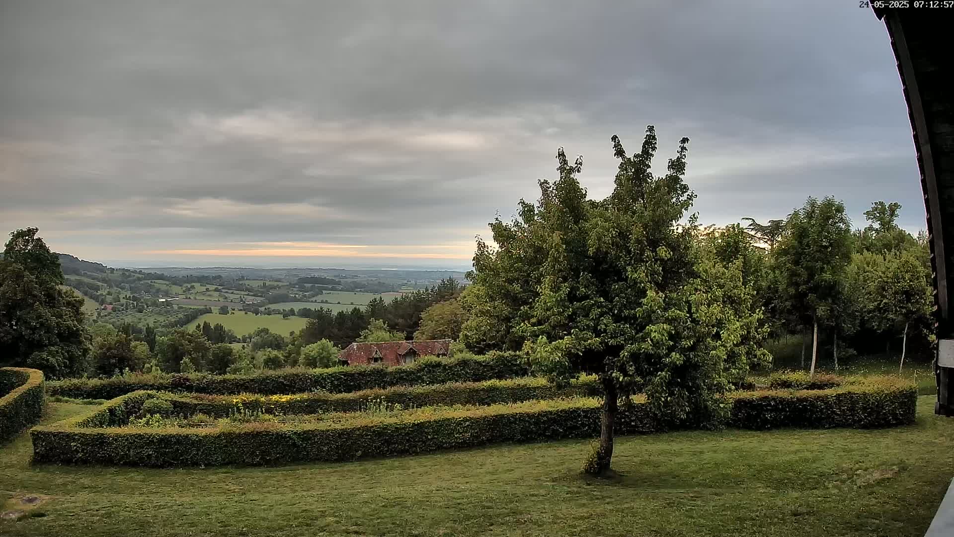 A sprawling green landscape with neatly trimmed hedges, a small building, and numerous trees under a cloudy sky, overlooks a distant valley.