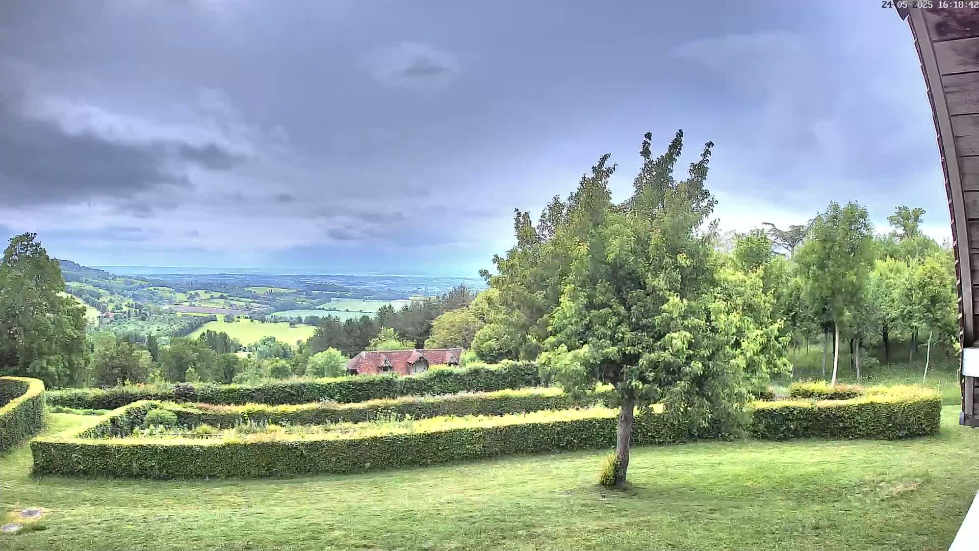 A verdant landscape under a cloudy sky features a manicured garden with hedges, a small building, and a distant view of rolling hills and a hazy horizon.