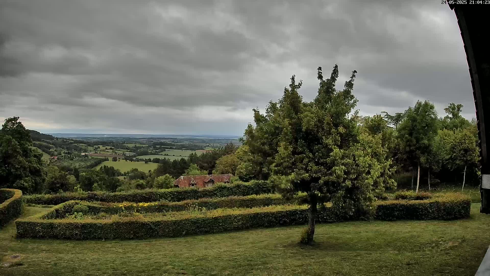 A verdant landscape with a neatly trimmed hedge surrounding a garden, overlooking a sprawling countryside under a cloudy sky.