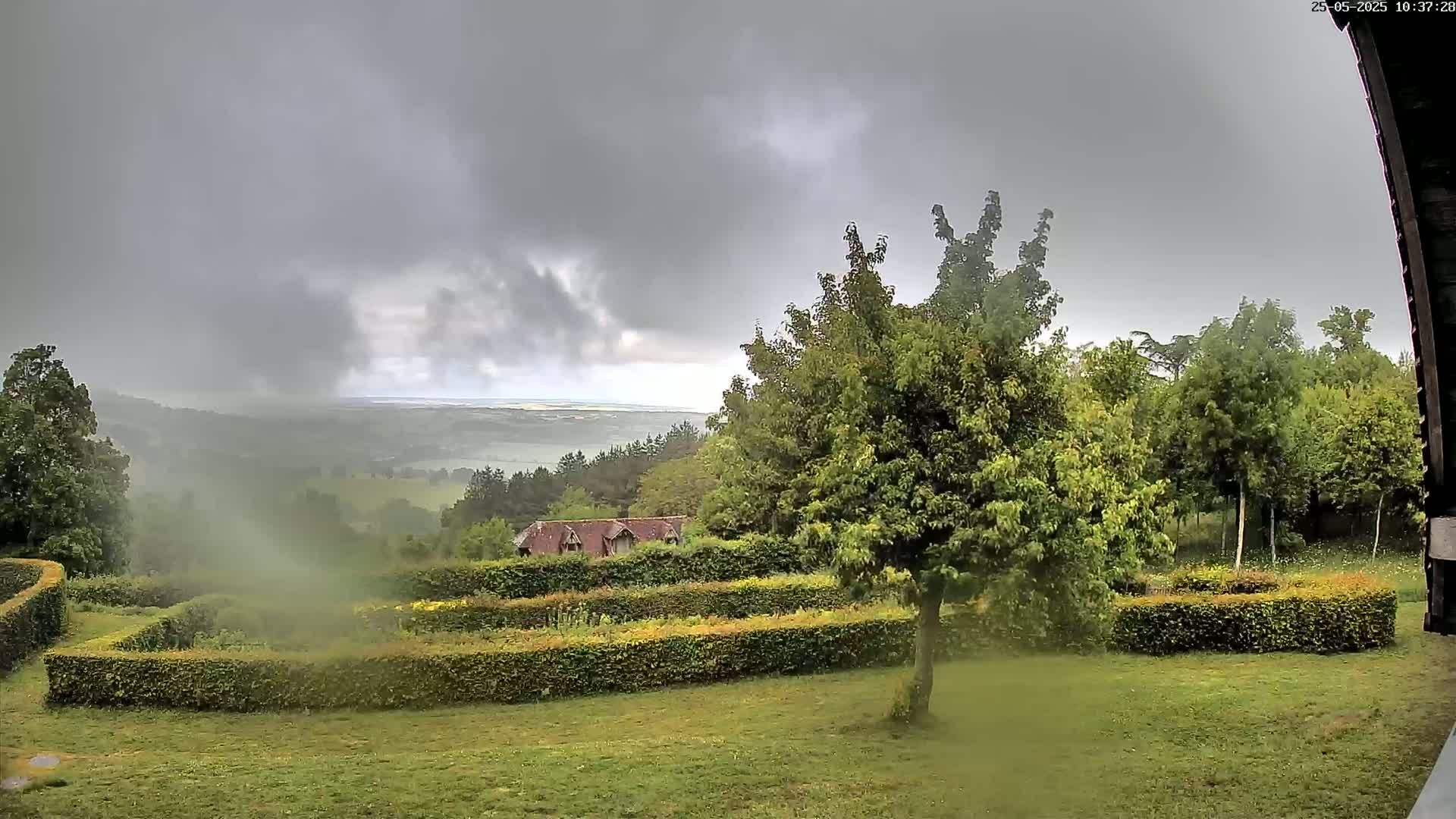 A view from a slightly elevated vantage point shows a verdant landscape with a geometrically-designed garden, trees, and a distant hazy horizon under an overcast sky.