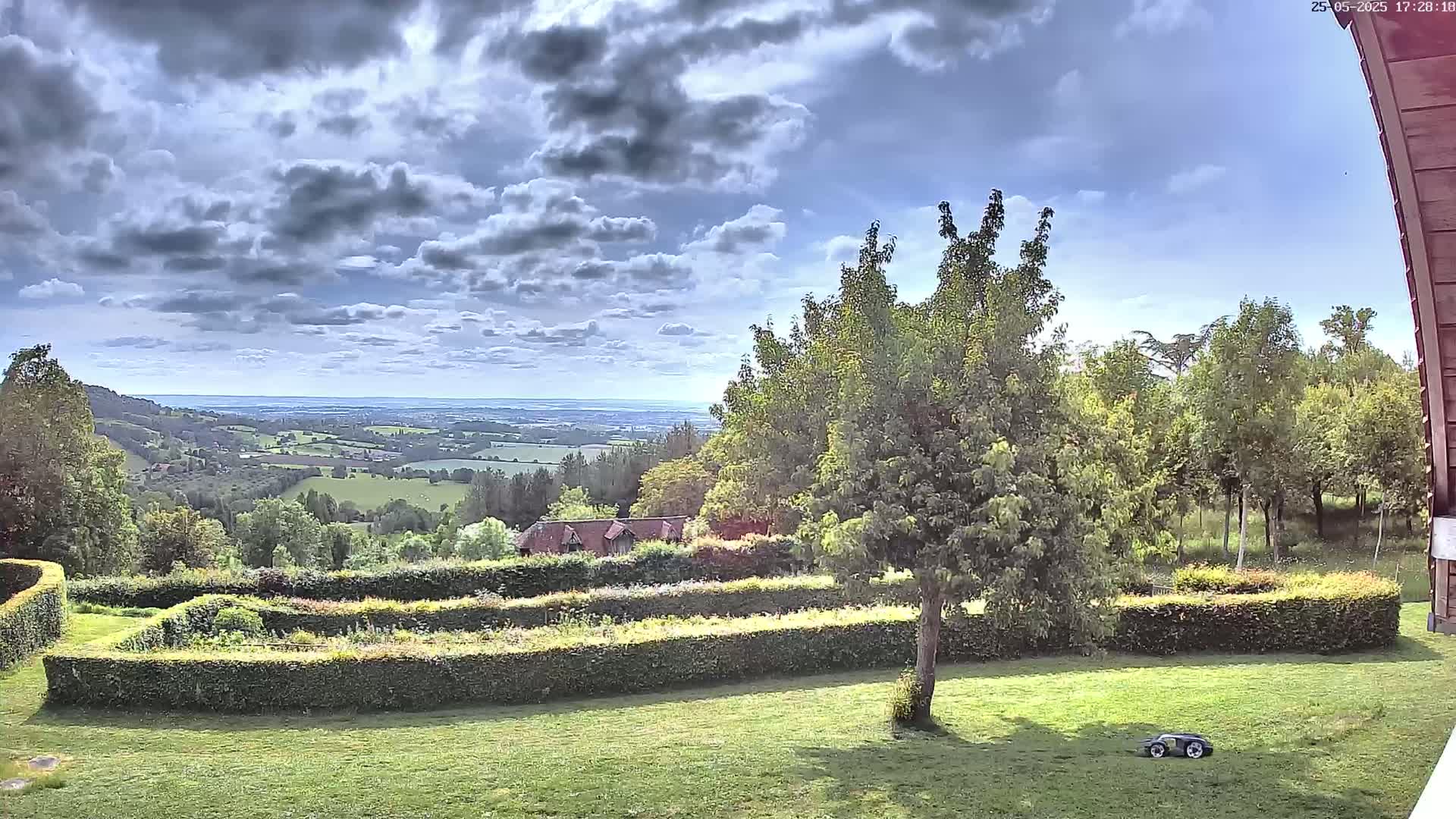 A partly cloudy sky overlooks a lush green landscape featuring a manicured lawn, hedges, and distant rolling hills, with a small robotic lawnmower visible in the foreground.
