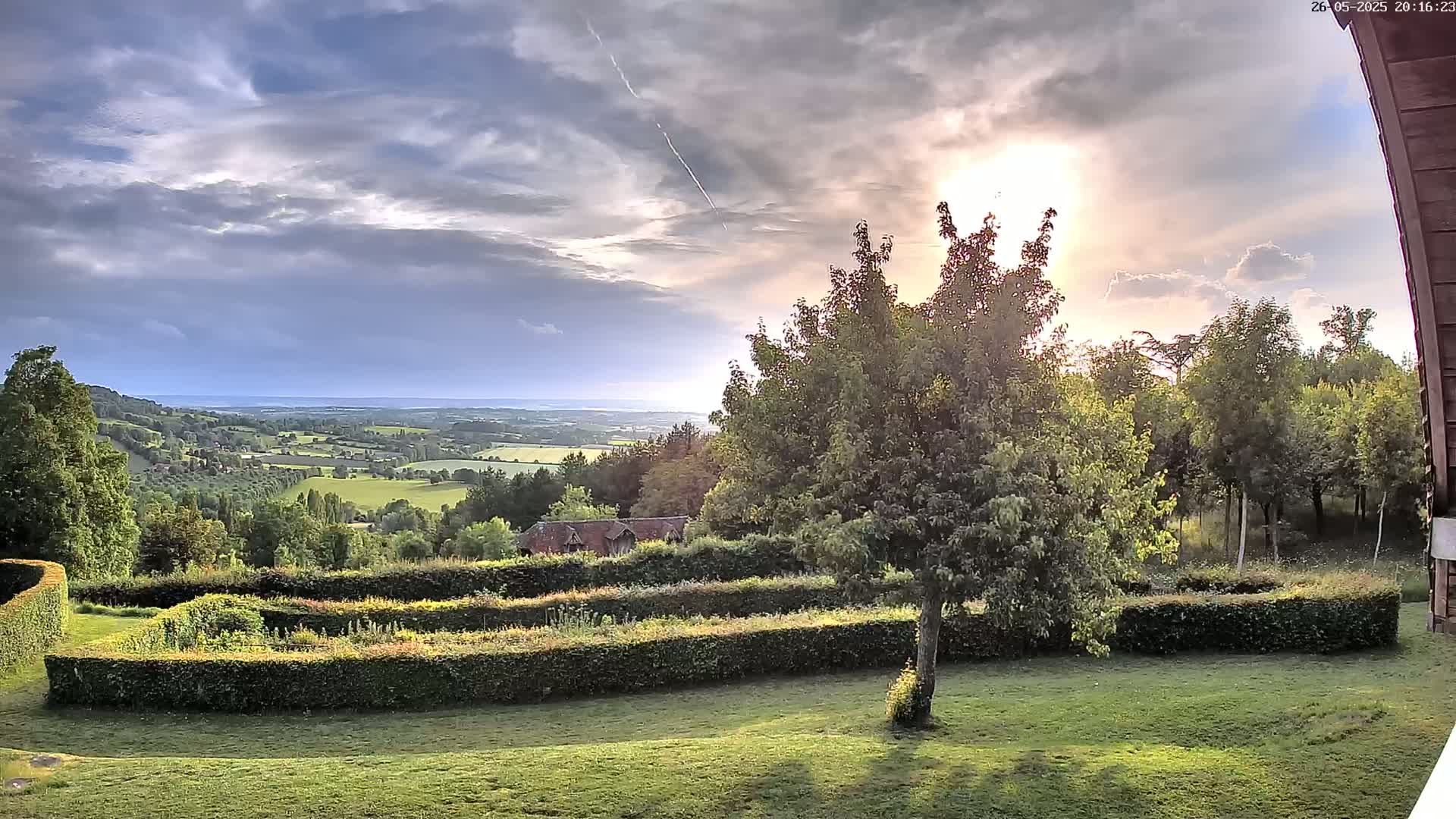 A partly cloudy sunset illuminates a verdant landscape featuring a manicured garden with hedges, trees, and a distant view of rolling hills and a body of water.