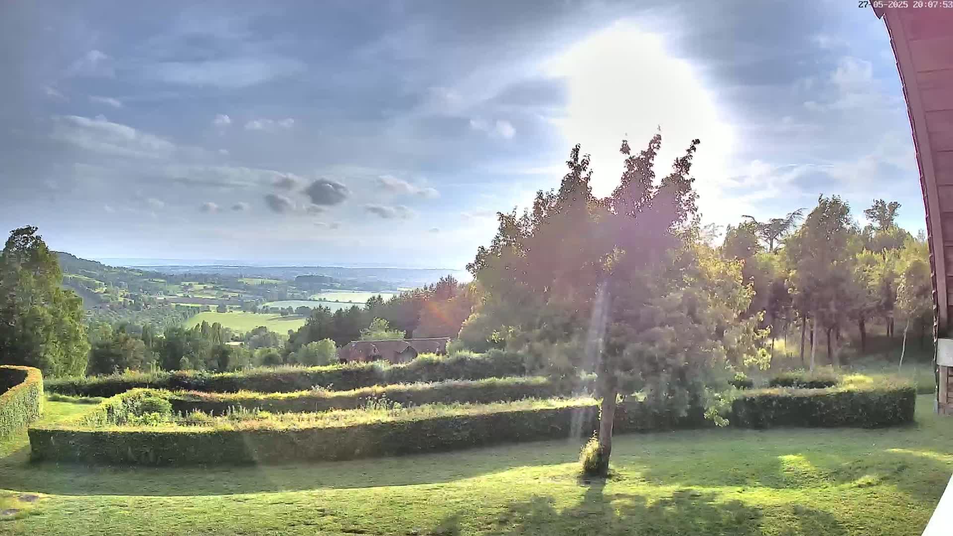 A partly sunny day reveals a picturesque landscape of rolling green hills, a distant body of water, and a manicured lawn with hedges, all viewed from an elevated position.