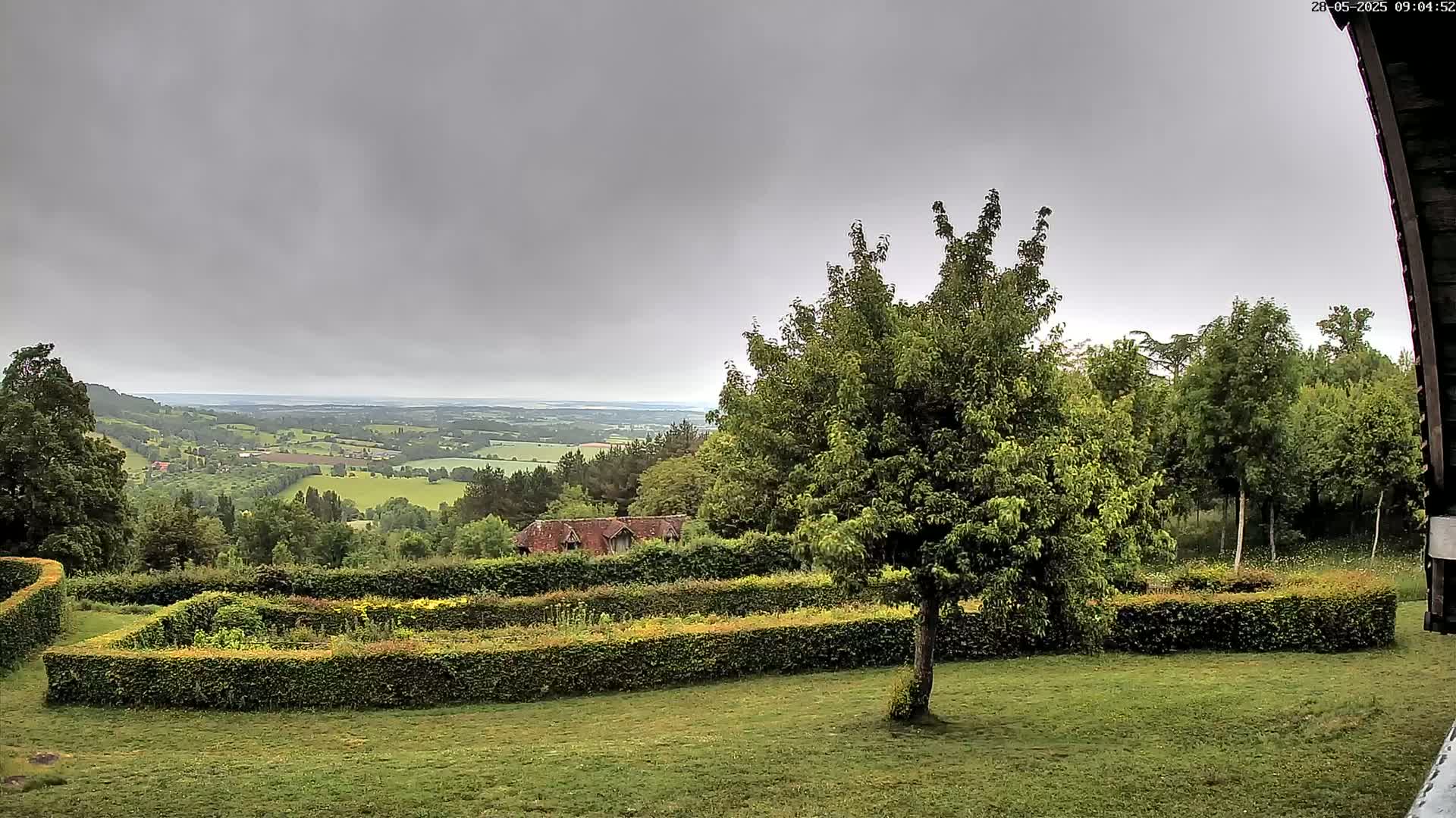 A verdant landscape under a gray sky reveals a meticulously manicured garden with hedges, trees, and a distant view of rolling hills.