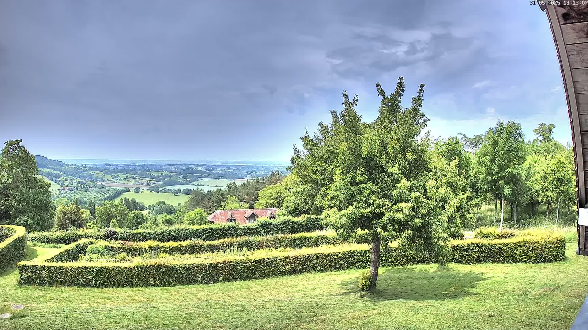 A verdant landscape featuring a neatly manicured garden with hedges, a tree, a distant building, and a rolling, expansive view under a partly cloudy sky.