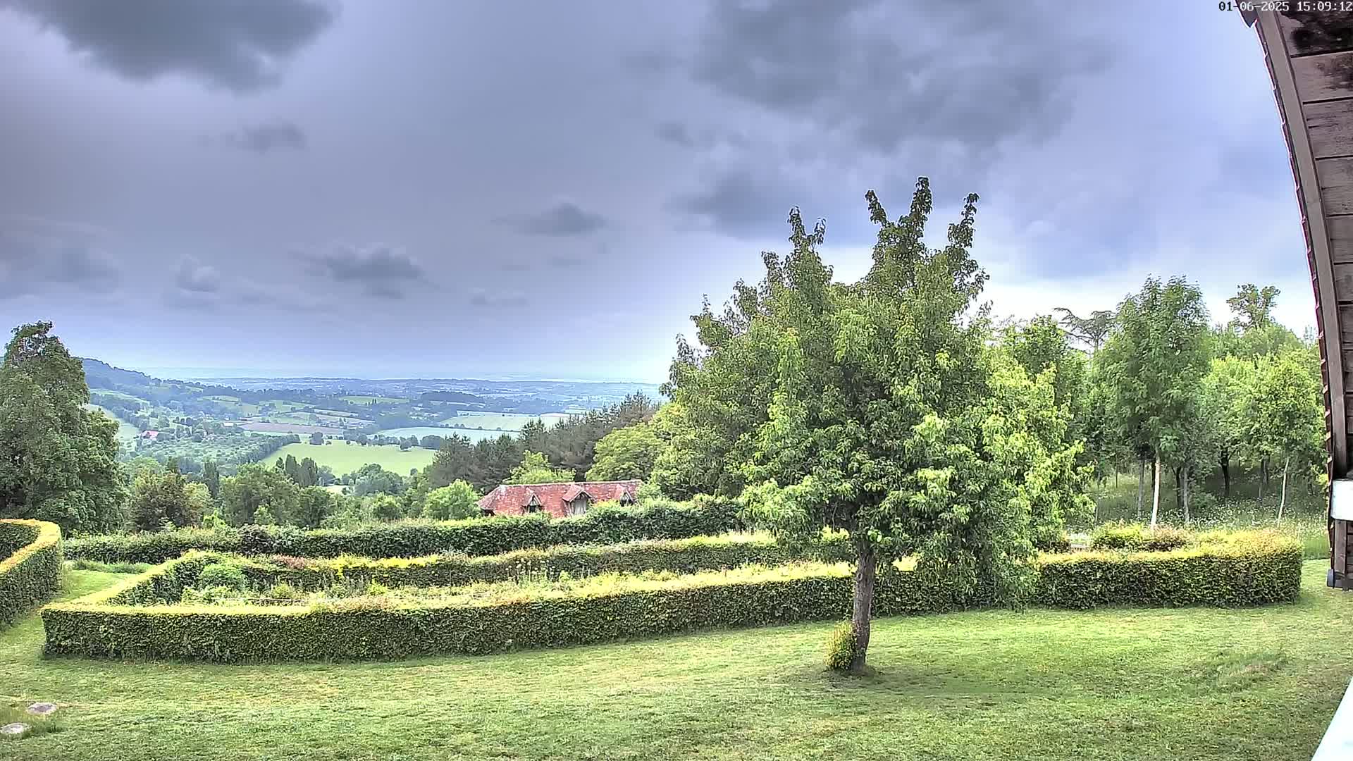 A verdant landscape with a meticulously manicured hedge-enclosed garden overlooks a rolling countryside under a cloudy sky.
