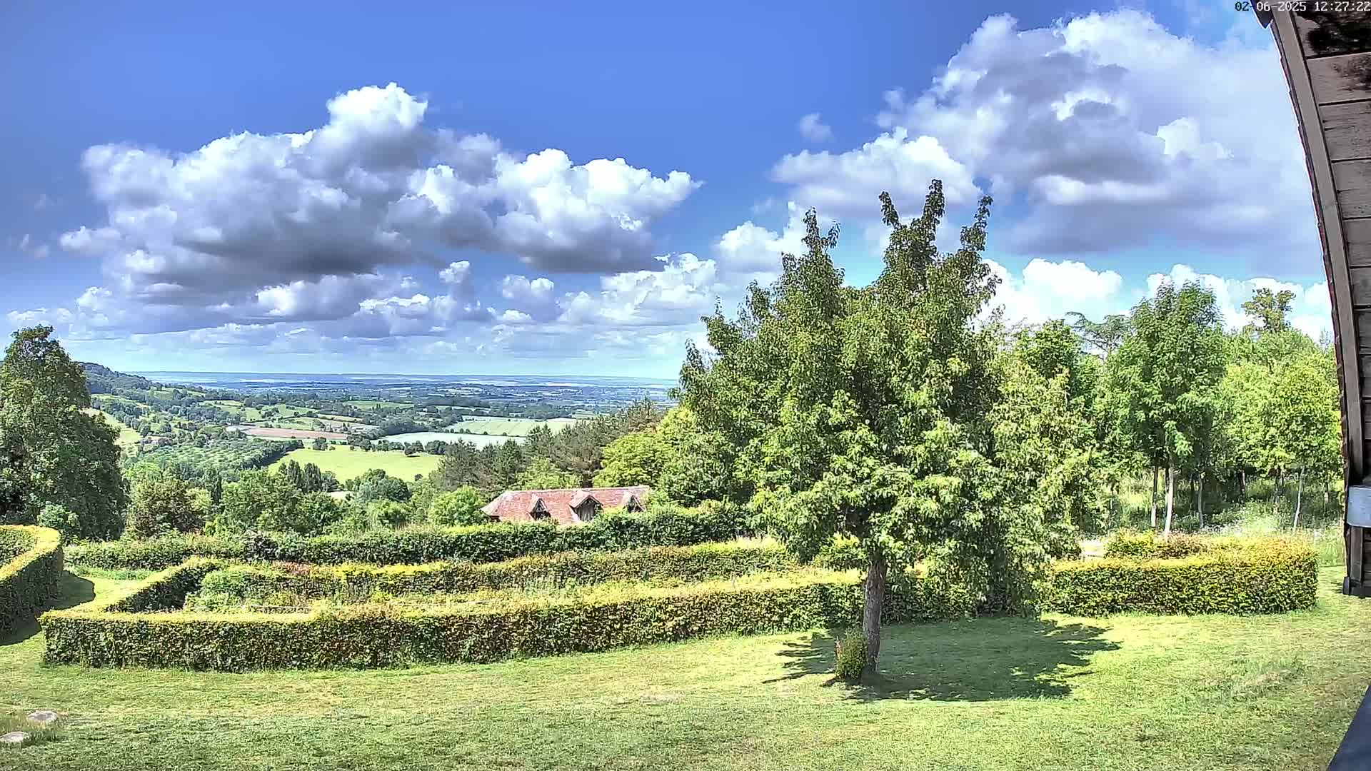 A sunny day with fluffy white clouds reveals a sprawling green landscape with a neatly trimmed hedge maze, a small building, and a large tree in the foreground, overlooking a distant valley.