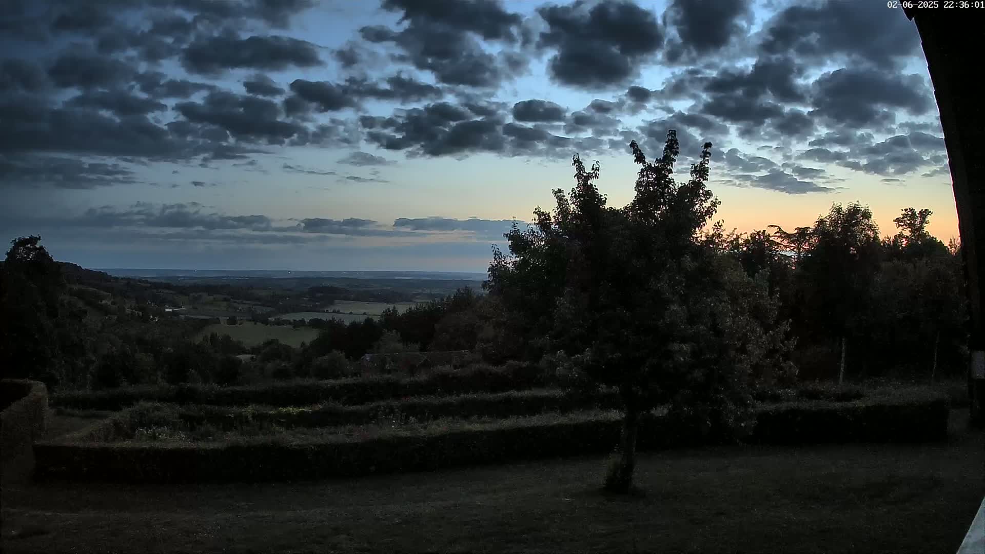 A dark, cloudy twilight sky overlooks a hilly landscape of dark green trees and shrubs, with a hint of orange on the horizon.